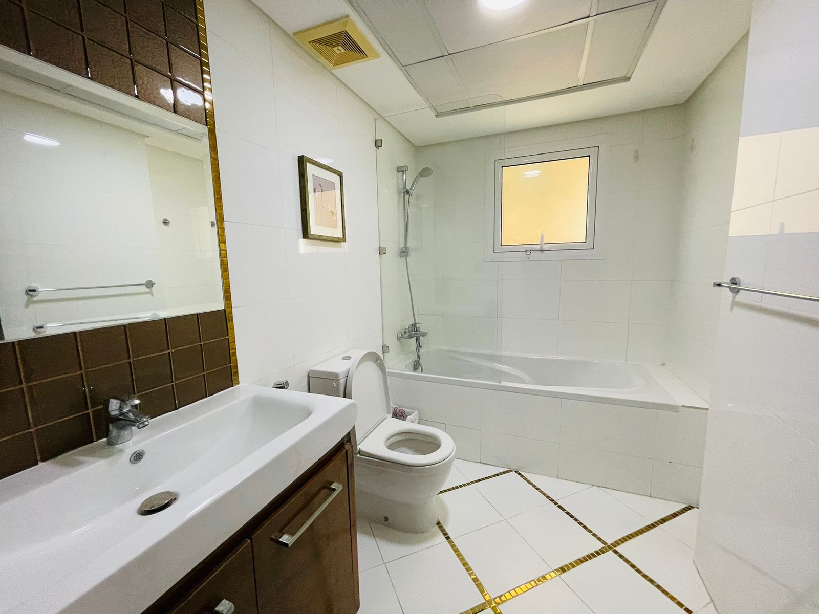 A clean, white tiled bathroom in a luxury apartment featuring a bathtub, toilet, sink, and two framed pictures on the wall, illuminated by natural light from a small window.