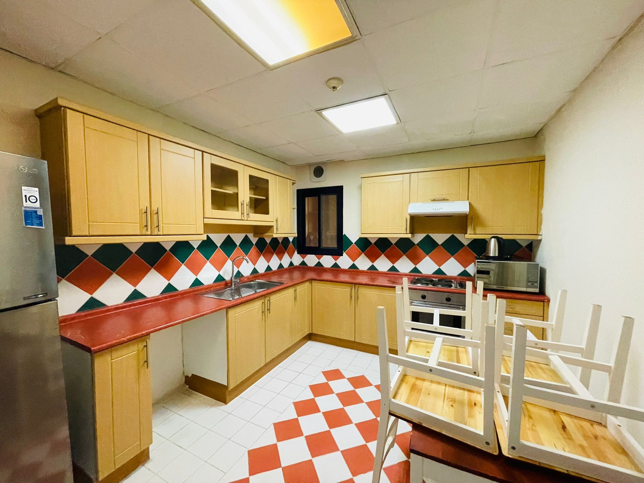 A brightly lit kitchen in a furnished apartment featuring wooden cabinets, a red countertop, patterned backsplash, and checkered floor tiles, with chairs inverted on a table to the right.