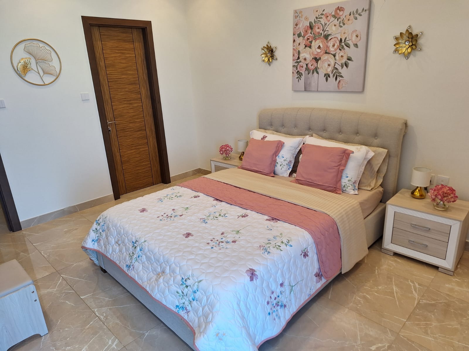 A neatly arranged bedroom in a luxury 1BR apartment in Juffair, featuring a floral-patterned bedspread, pink pillows, and decorative wall art.