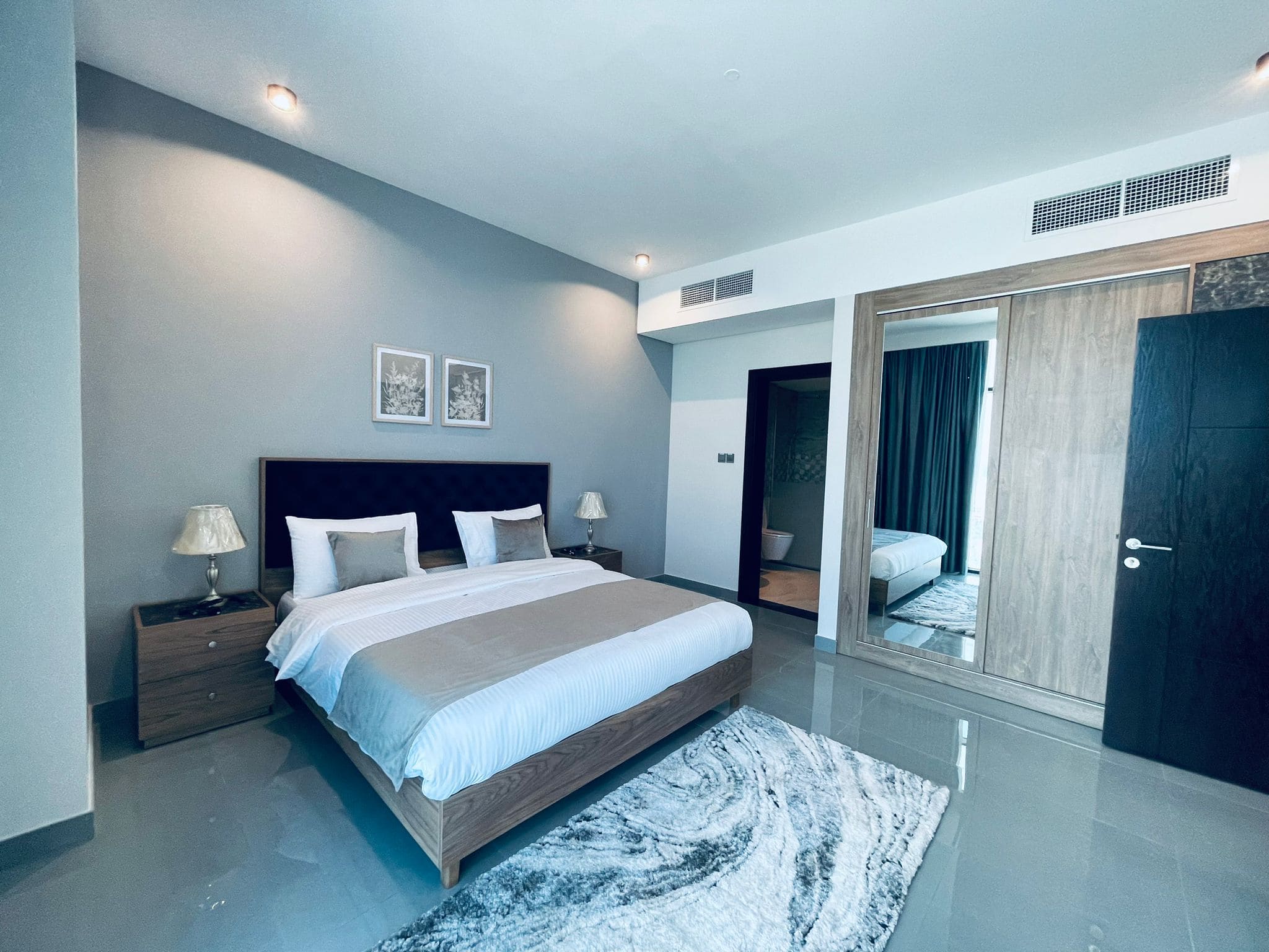 Modern bedroom with a large bed, white bedding, two bedside lamps, a gray rug, and an open door leading to another room.