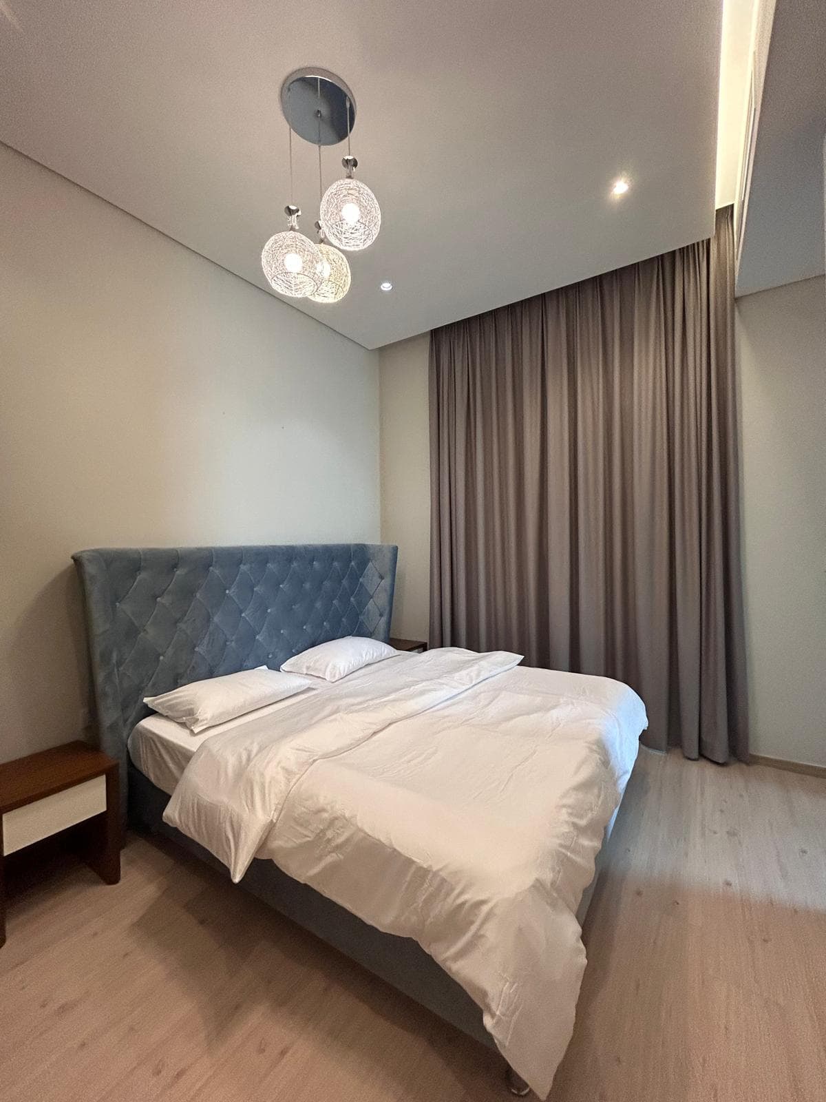 Modern bedroom in a furnished apartment, with an unmade bed, gray upholstered headboard, and elegant pendant lights, featuring a large, draped window.