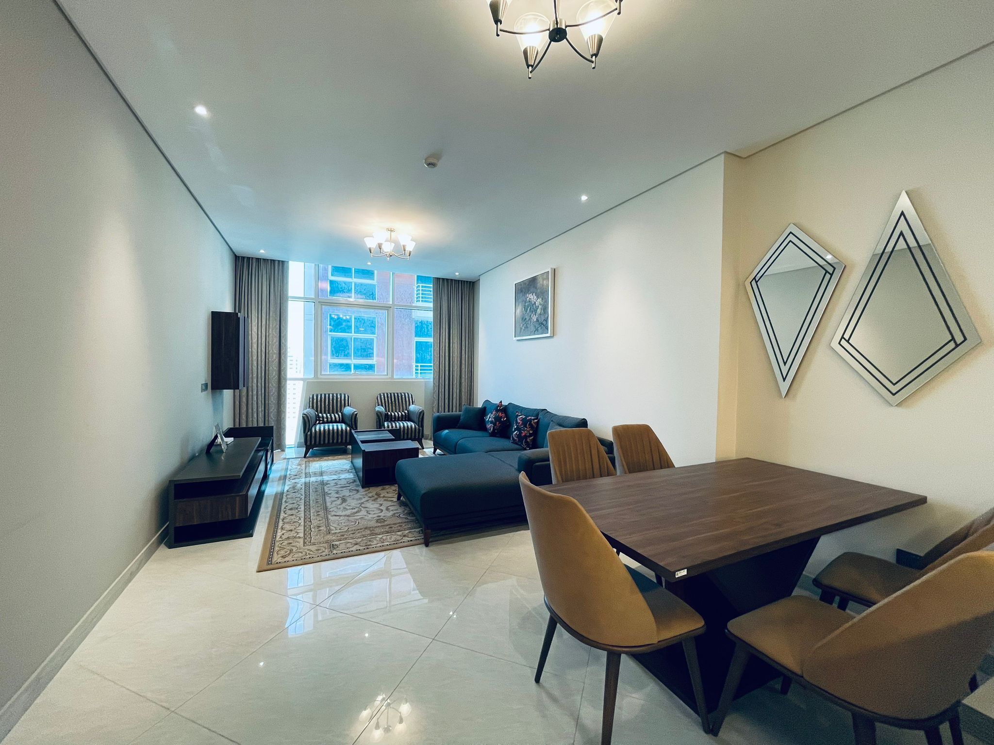 Luxury 1BR apartment in Juffair with a modern living room featuring a blue sofa, striped chairs, and a wooden dining table, lit by ceiling lights and adorned with decorative wall art.