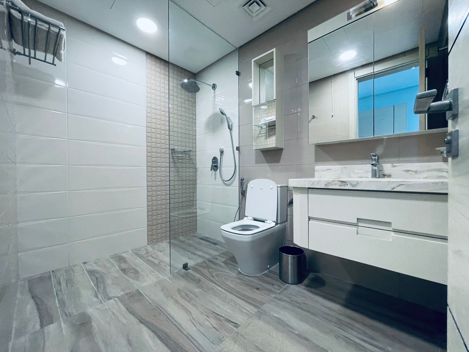 Luxury 1BR Apartment in Juffair features a modern bathroom interior with a glass shower stall, white toilet, and a vanity with a sink and mirror. Light tiles and wood flooring complete the