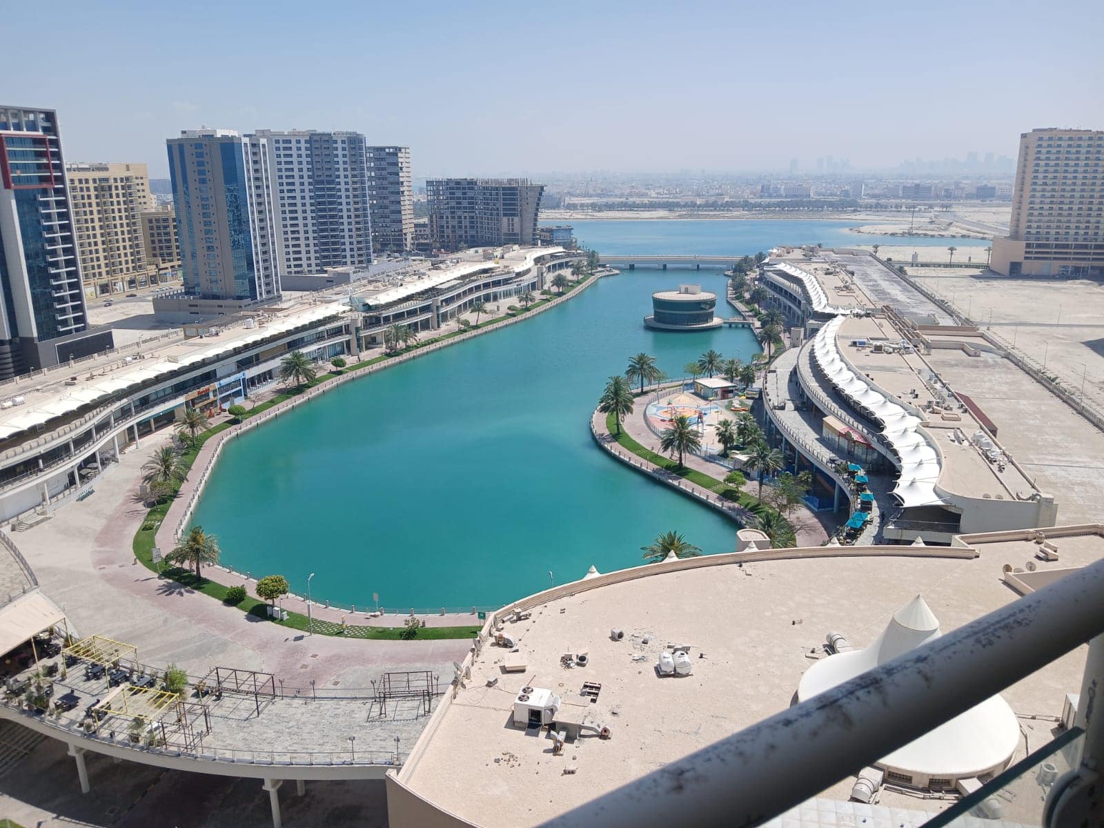 Aerial view of a vibrant waterfront promenade in Amwaj with high-rise buildings, a turquoise artificial lake, and a city skyline in the distance.