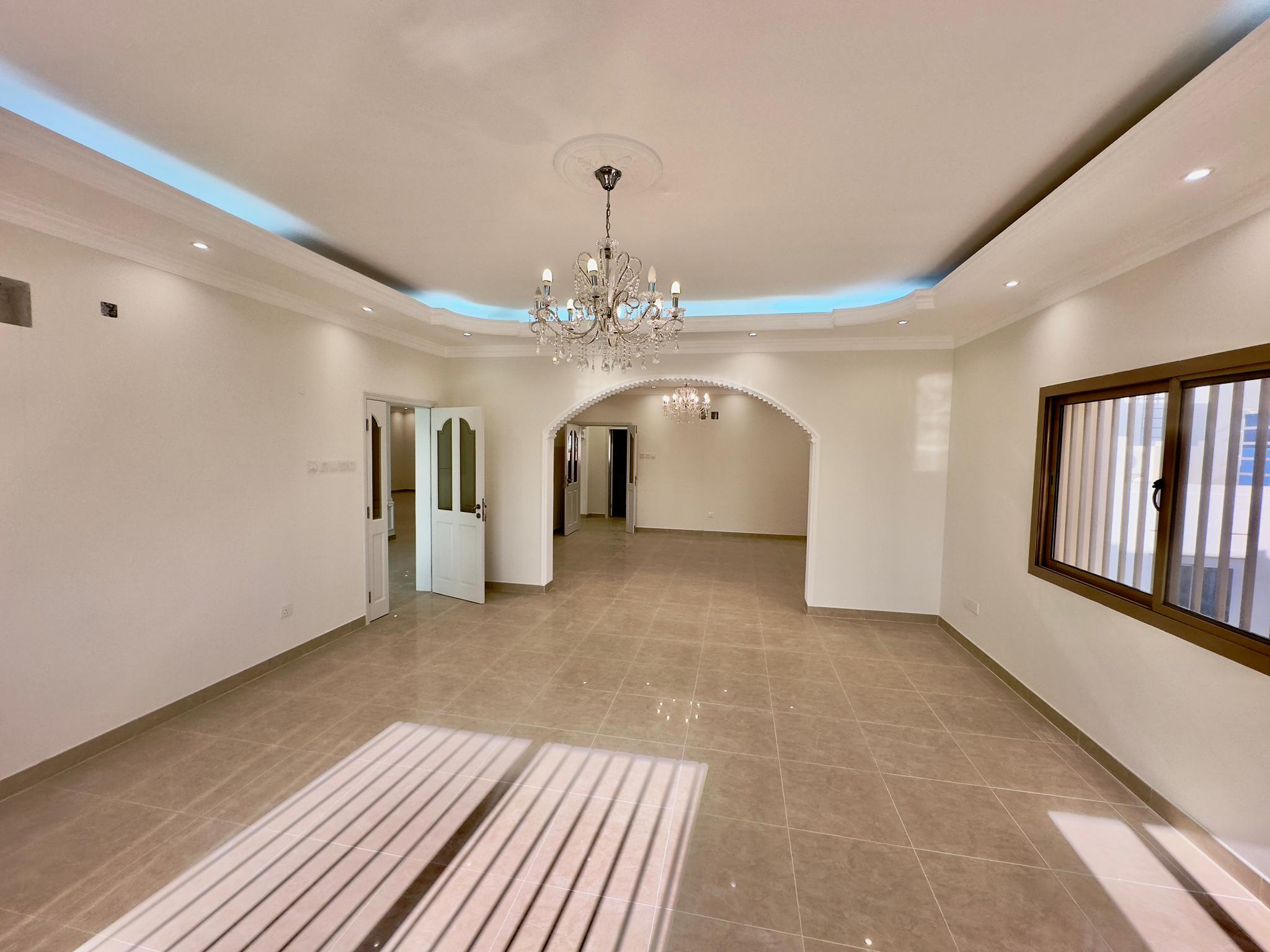 Spacious, empty living room with tiled floors, arched wall openings, recessed ceiling with blue led lights, and a crystal chandelier.