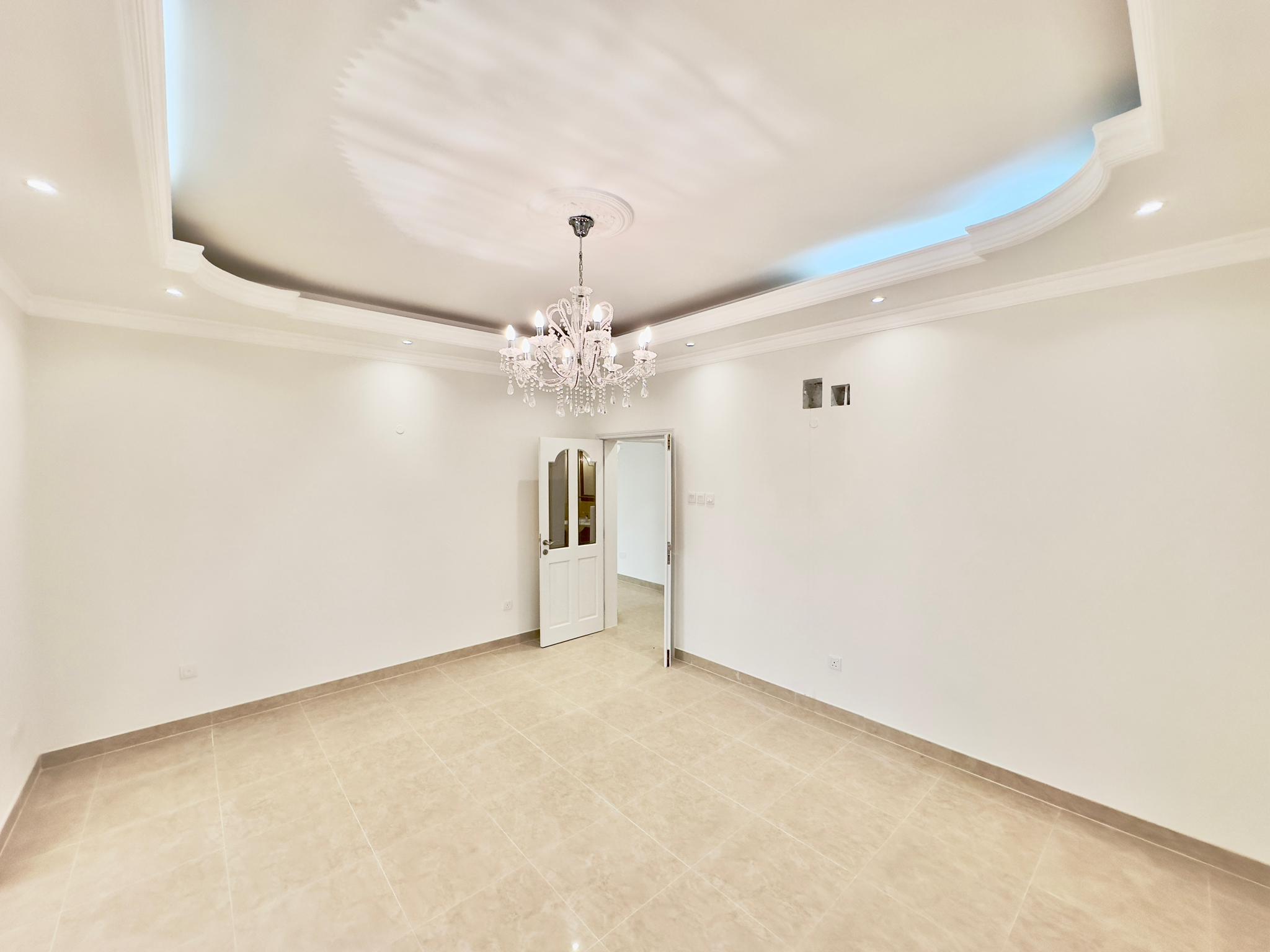 Spacious, empty room in a luxury apartment with white walls, coved ceiling with blue lighting, and a crystal chandelier. Tile flooring is visible and doors lead to other areas.