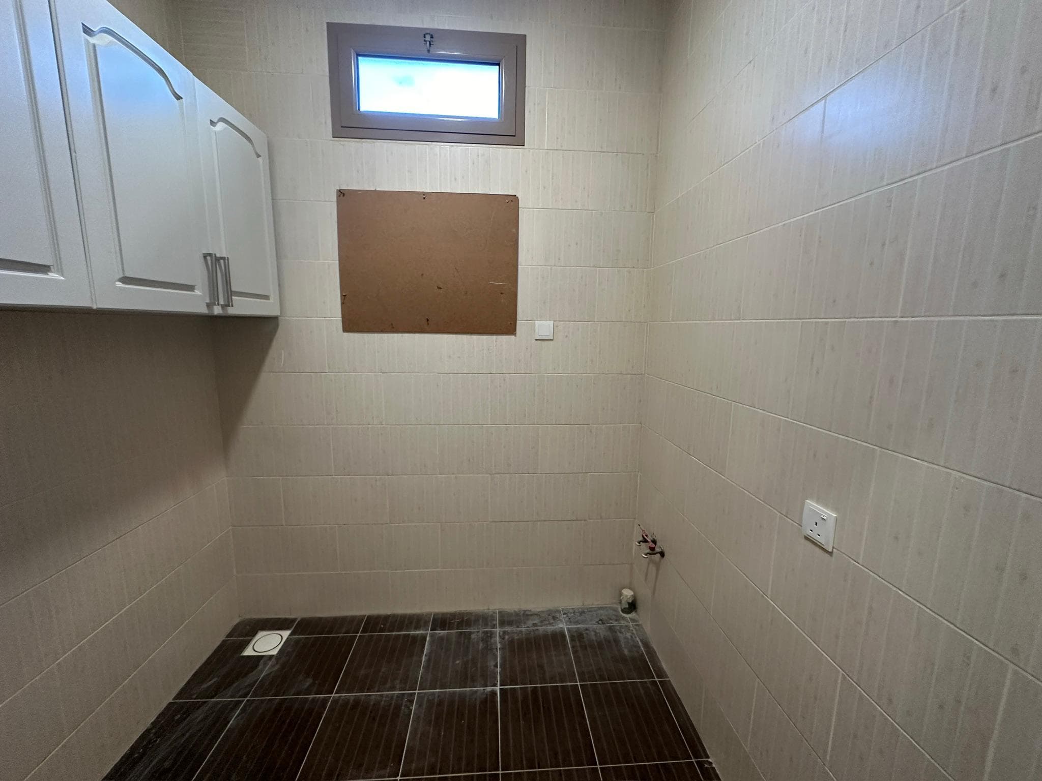 A small, partially tiled bathroom with a squat toilet, a hanging cabinet, and an empty board on the wall under a small window.