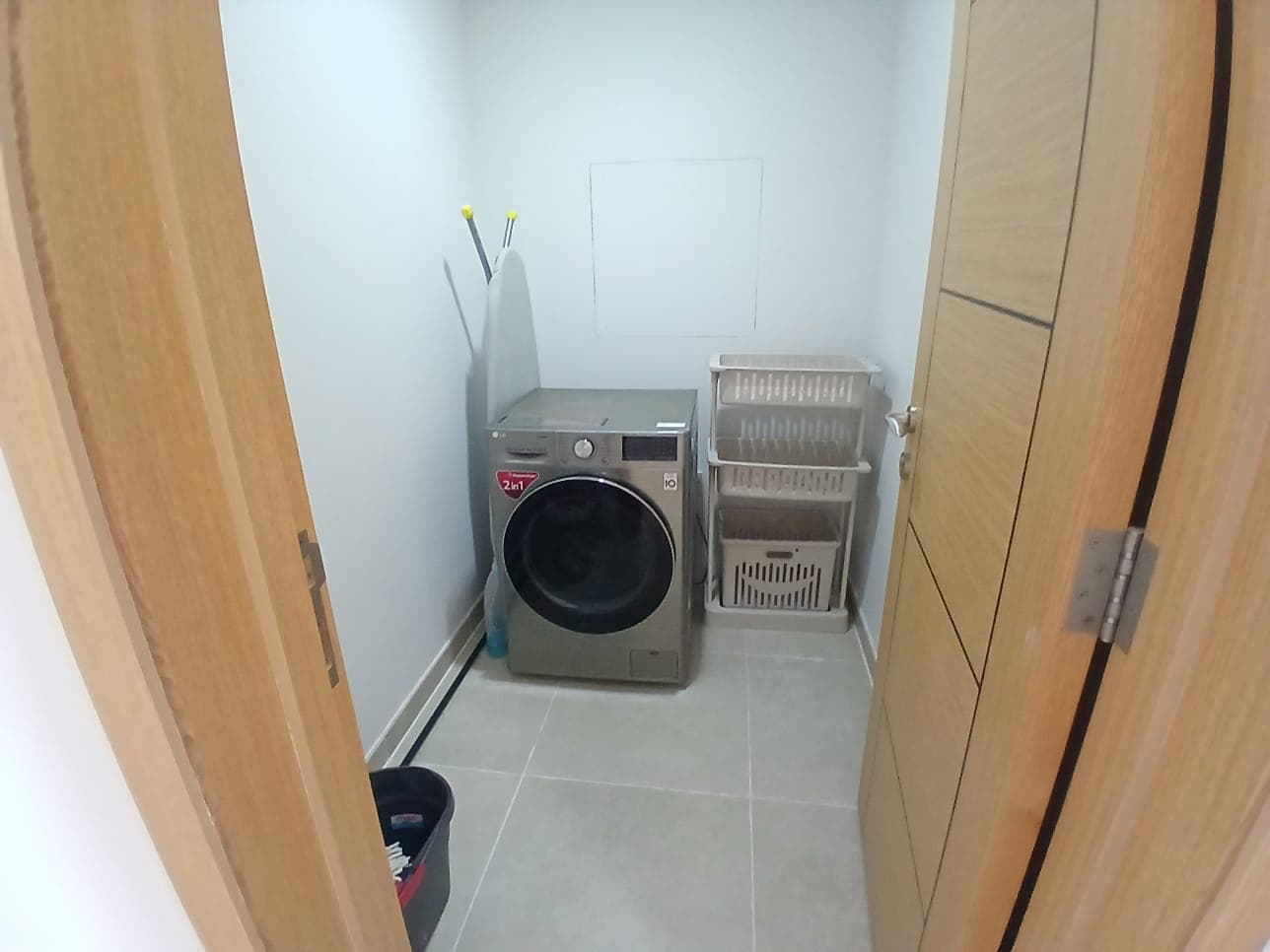 A small laundry room in an Amwaj flat for rent, featuring a washing machine, a wall-mounted drying rack, and cleaning supplies, viewed from an open door.