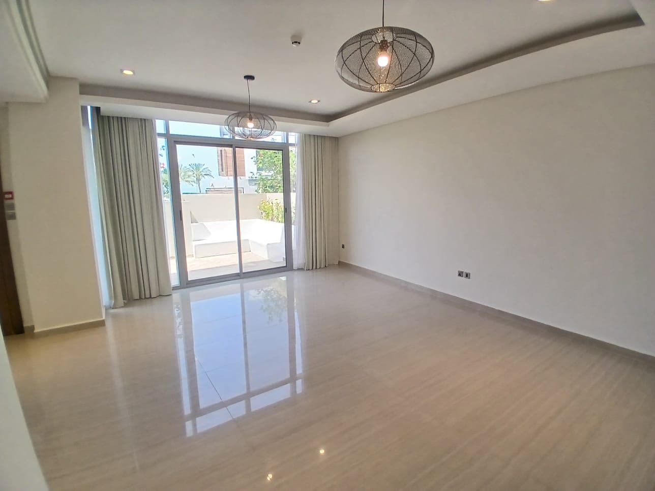 Bright, empty room in a modern villa for rent with glossy tiled floor, large windows with curtains leading to a balcony, and modern hanging lights.