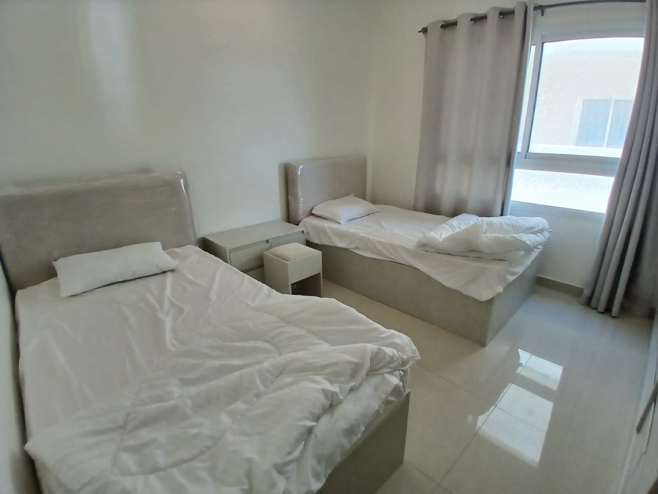 A simple modern bedroom in a balcony apartment with two single beds covered in white linens, a gray upholstered headboard, and a window with light curtains.