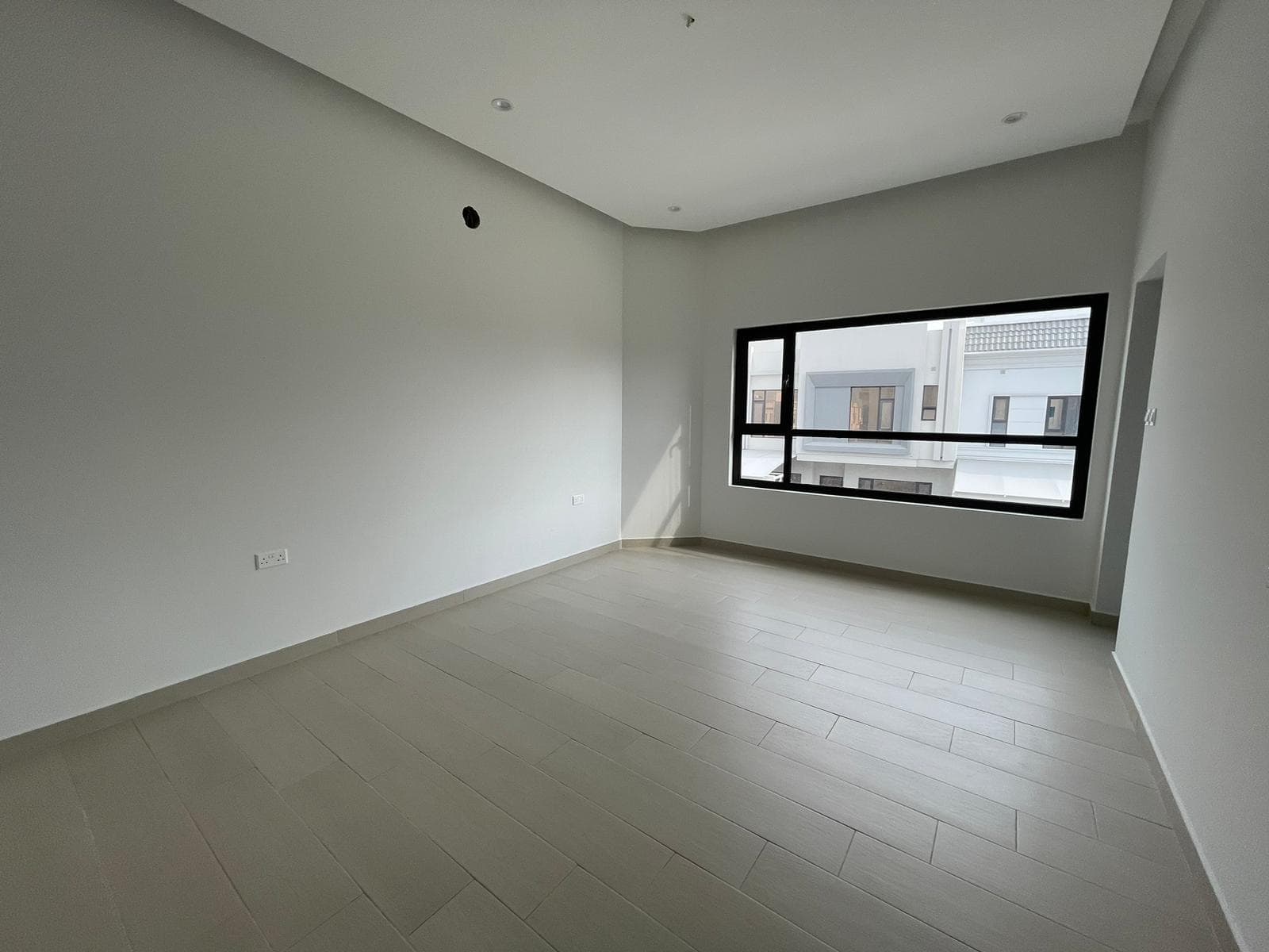 Empty living room with white walls and ceramic tiled flooring in a villa for sale, featuring a large, rectangular window showing a view of neighboring houses in Bani Jamra.