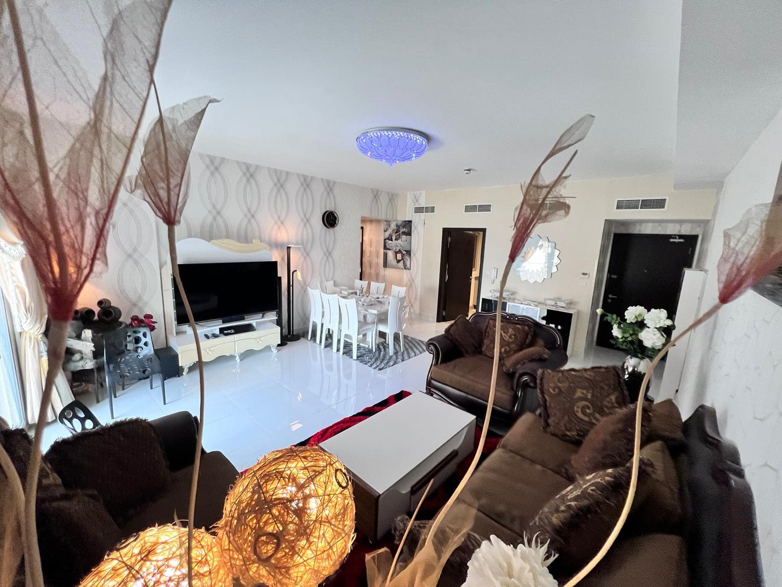 A spacious living room in a luxury apartment in Juffair, featuring a brown sofa set, a white coffee table, a large TV, and a dining table with white chairs. Modern decor includes plants and artistic lighting fixtures. This elegant space is now available for sale.