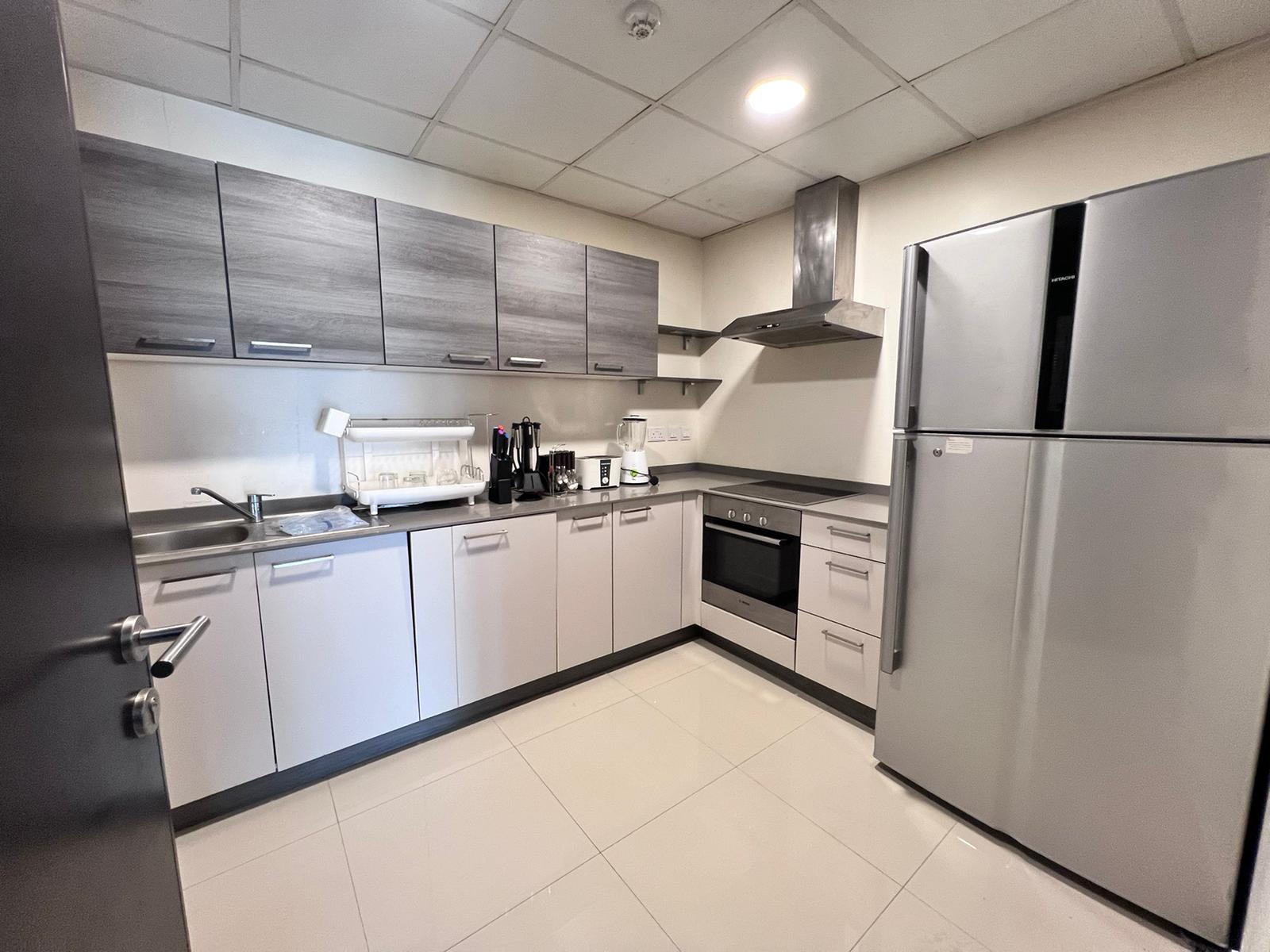 Modern kitchen with light grey cabinets, stainless steel appliances, including a fridge, oven, microwave, and stovetop. The counter has a sink, knife set, toaster, and kettle with white tile flooring. This luxury apartment in Juffair is now on sale!