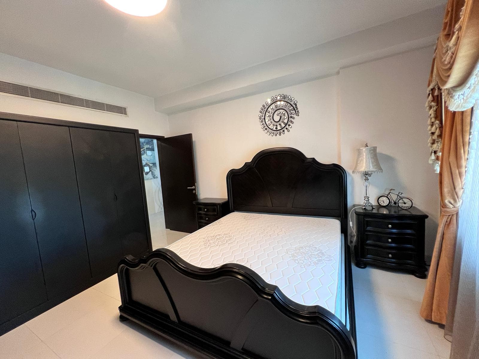 In the luxury apartment located in Juffair, the bedroom features a dark wood bed frame, matching bedside tables, and a black wardrobe. A decorative wall piece hangs above the bed, while heavy curtains frame the window. This elegant living space is currently on sale.