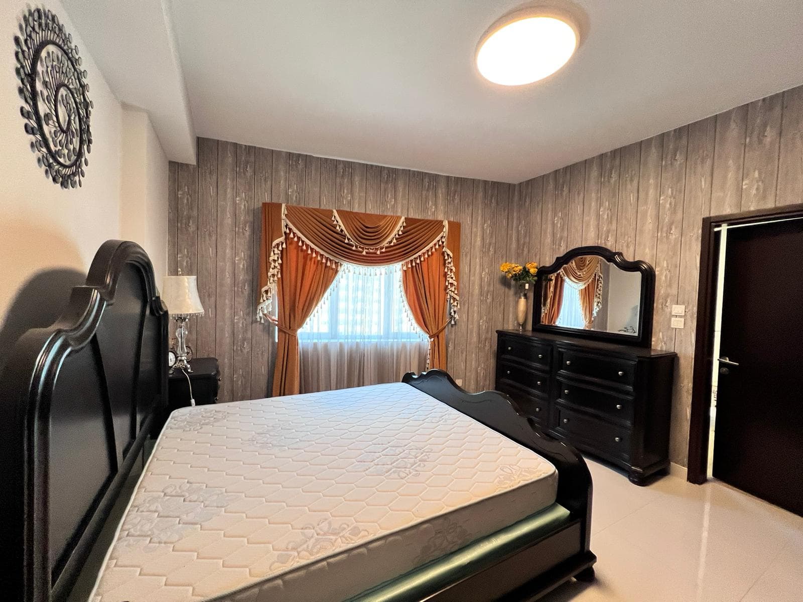 Bedroom in a luxury apartment in Juffair, featuring dark wooden furniture including a bed frame, dresser, and mirror. The bed has a bare mattress. The window is adorned with orange and white drapes with a valance. The walls are covered in textured wallpaper. Available for sale now.