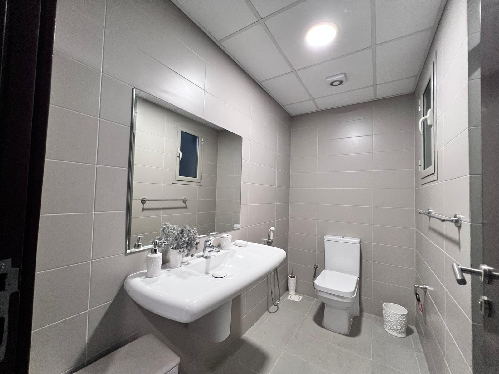 A modern bathroom in a luxury apartment for sale in Juffair, featuring grey tile walls and flooring. It includes a white sink with toiletries, a large mirror, a toilet, and a window. The room is well-lit by a ceiling light.