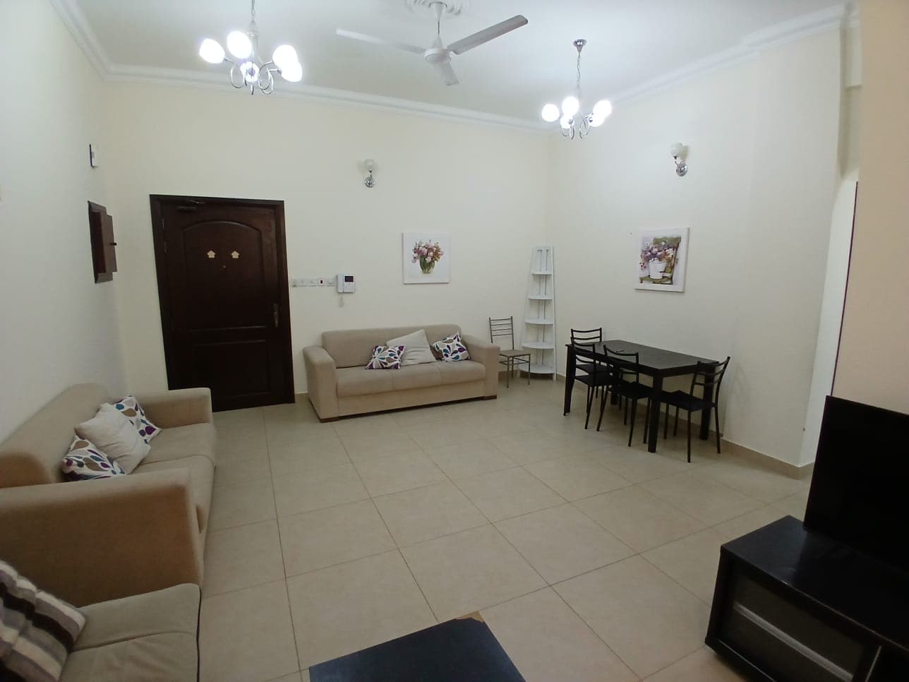 A furnished living room with a ceiling fan and two light fixtures. It has two sofas, a dining table with four chairs, wall art, and a closet. The room features a tiled floor and a wall-mounted TV.