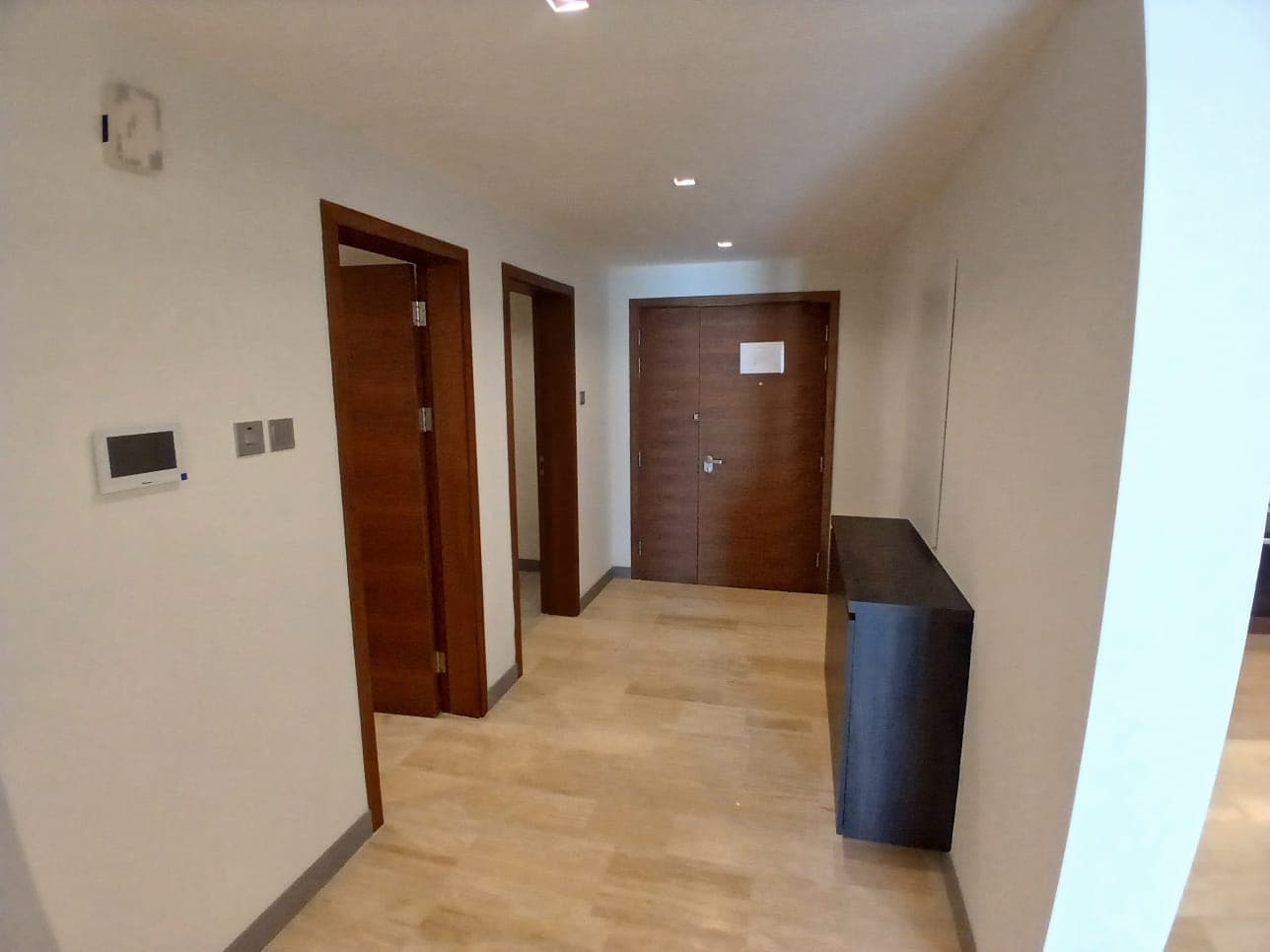 A hallway with wooden doors on the left, a thermostat on the wall, and a dark-colored cabinet on the right side. This flat's hallway leads to a main door at the end, just steps away from experiencing life on Dilmunia Island.