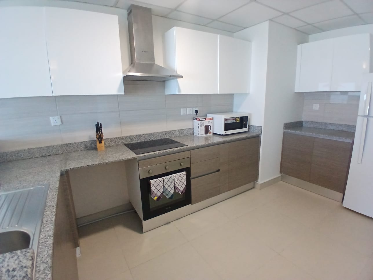 Modern kitchen in a furnished flat on Amwaj Islands features white and wooden cabinets, stainless steel appliances including a stove, microwave, and refrigerator, with a grey countertop and a knife set on the counter.