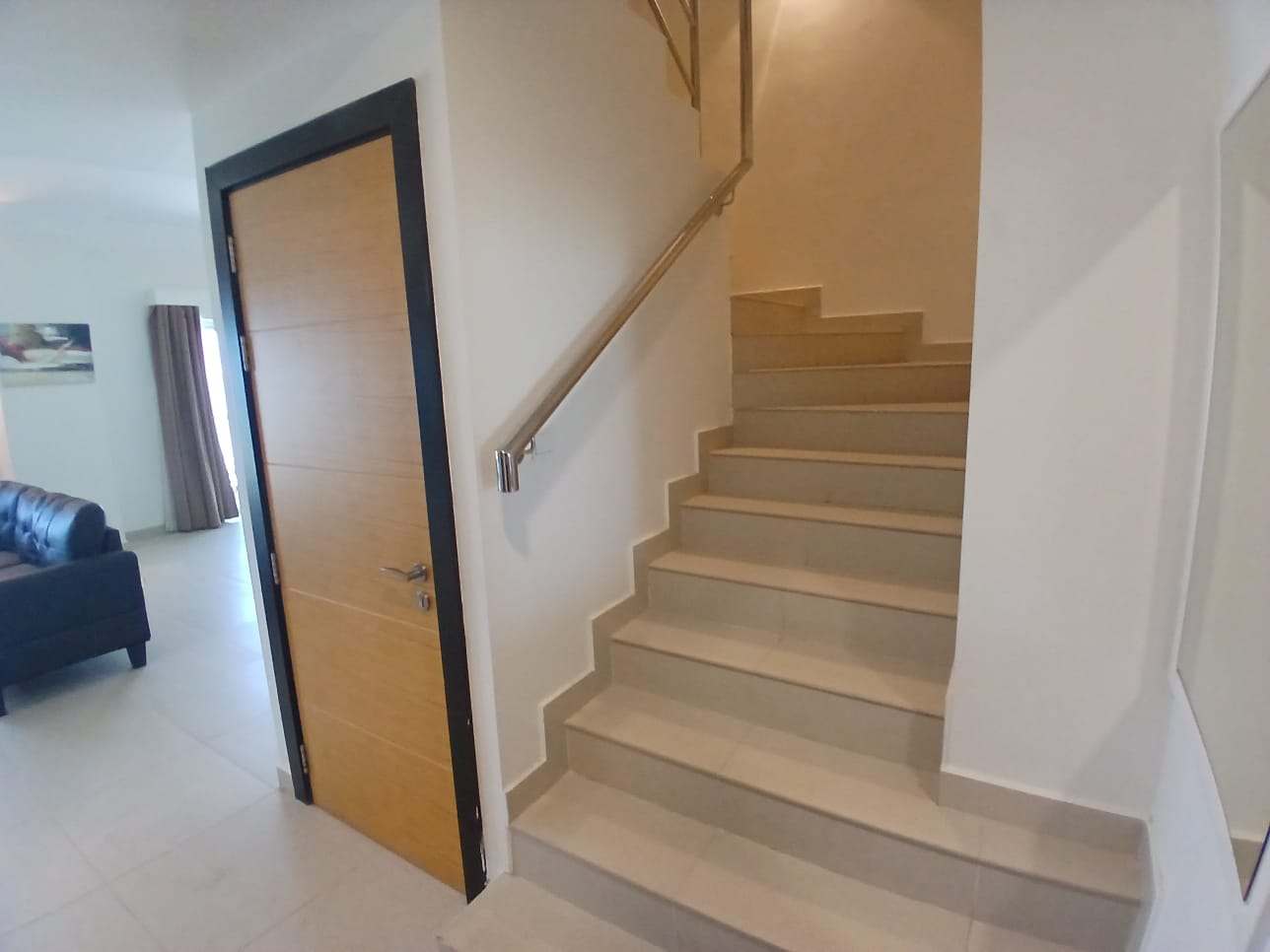 Indoor space with a wooden door on the left, a staircase with a metal handrail leading upwards on the right, and a view of the fully furnished flat's living room with a couch in the background in Amwaj Islands.