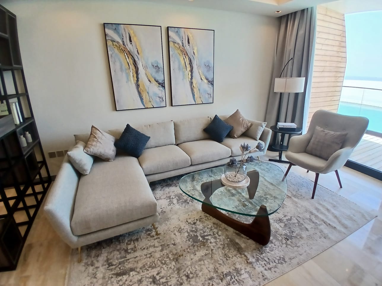 A modern living room in a flat for sale features a grey L-shaped sofa, a glass coffee table, an armchair, two abstract paintings, and a floor lamp. A large window offers a view of the Dilmunia balcony and the sea.