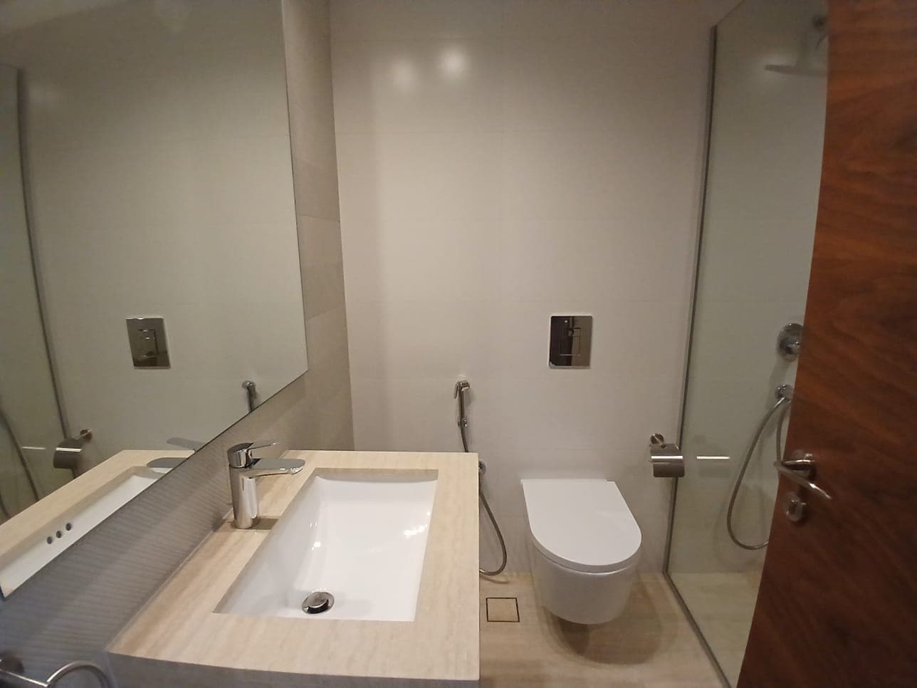 A modern bathroom in a flat for sale at Dilmunia features a wall-mounted toilet and a square sink on a beige countertop. A large mirror reflects the shower area with its glass partition, while a wooden door is partly visible, adding to the property's contemporary appeal.