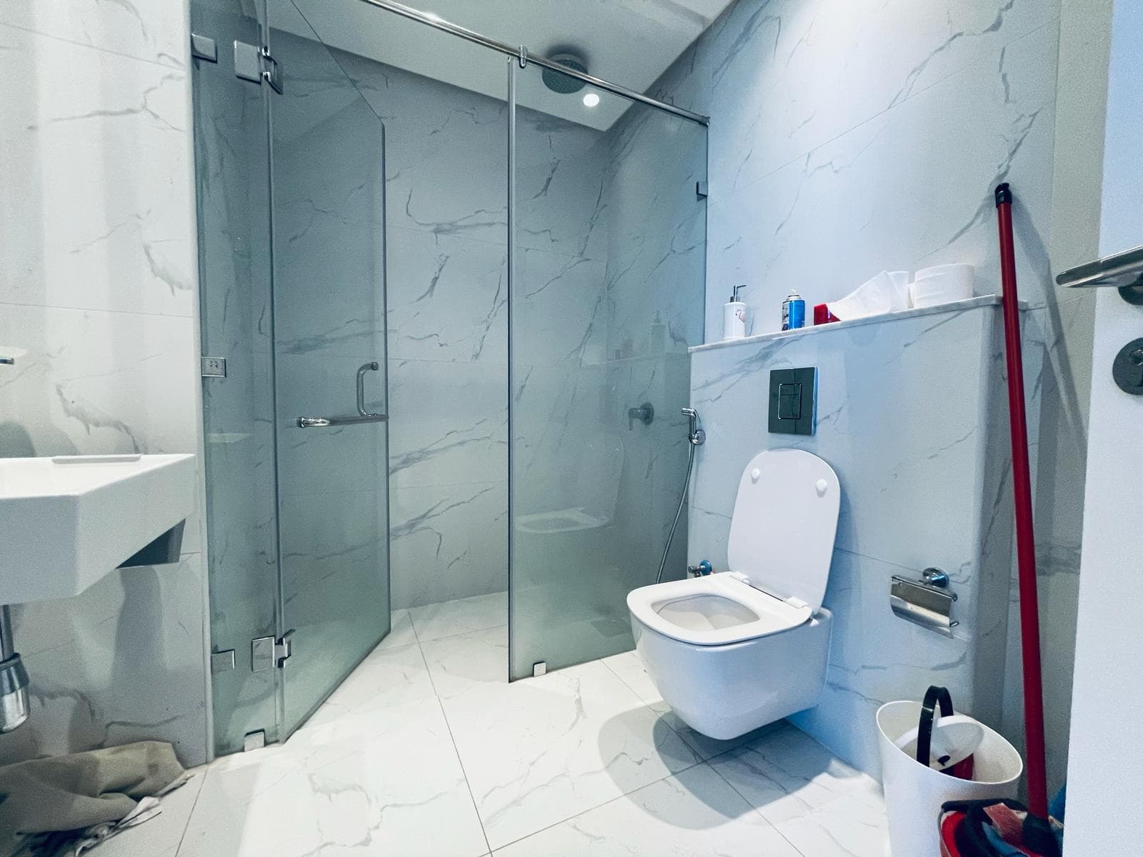 A modern bathroom in an amazing studio for rent in the Seef area, featuring marble walls and flooring, a glass shower enclosure, a white toilet, a wall-mounted sink, and various toiletries on a shelf. The scene also includes a mop and bucket.