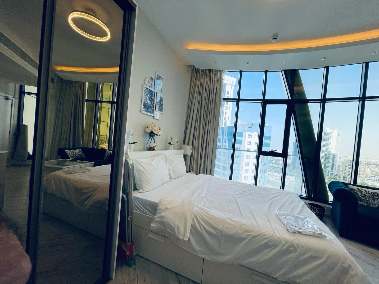 A modern studio for rent in the Seef area features a neatly made bed, large floor-to-ceiling windows, a wall mirror, a small sofa, and stunning cityscape views.