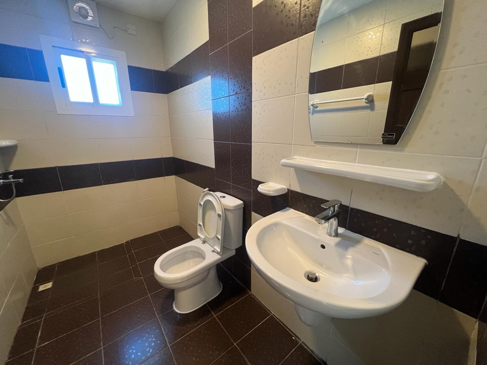 A modern bathroom in a rental property, featuring a toilet, wall-mounted sink, mirror, tiled walls, and small window with blinds.