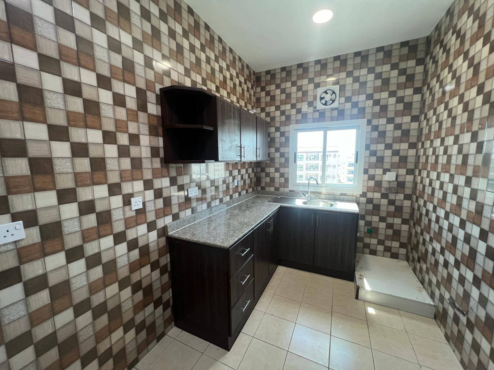 Modern kitchen with checkered brown and beige tiles, dark wood cabinets, granite countertops, and a window with a city view in the Hidd area.