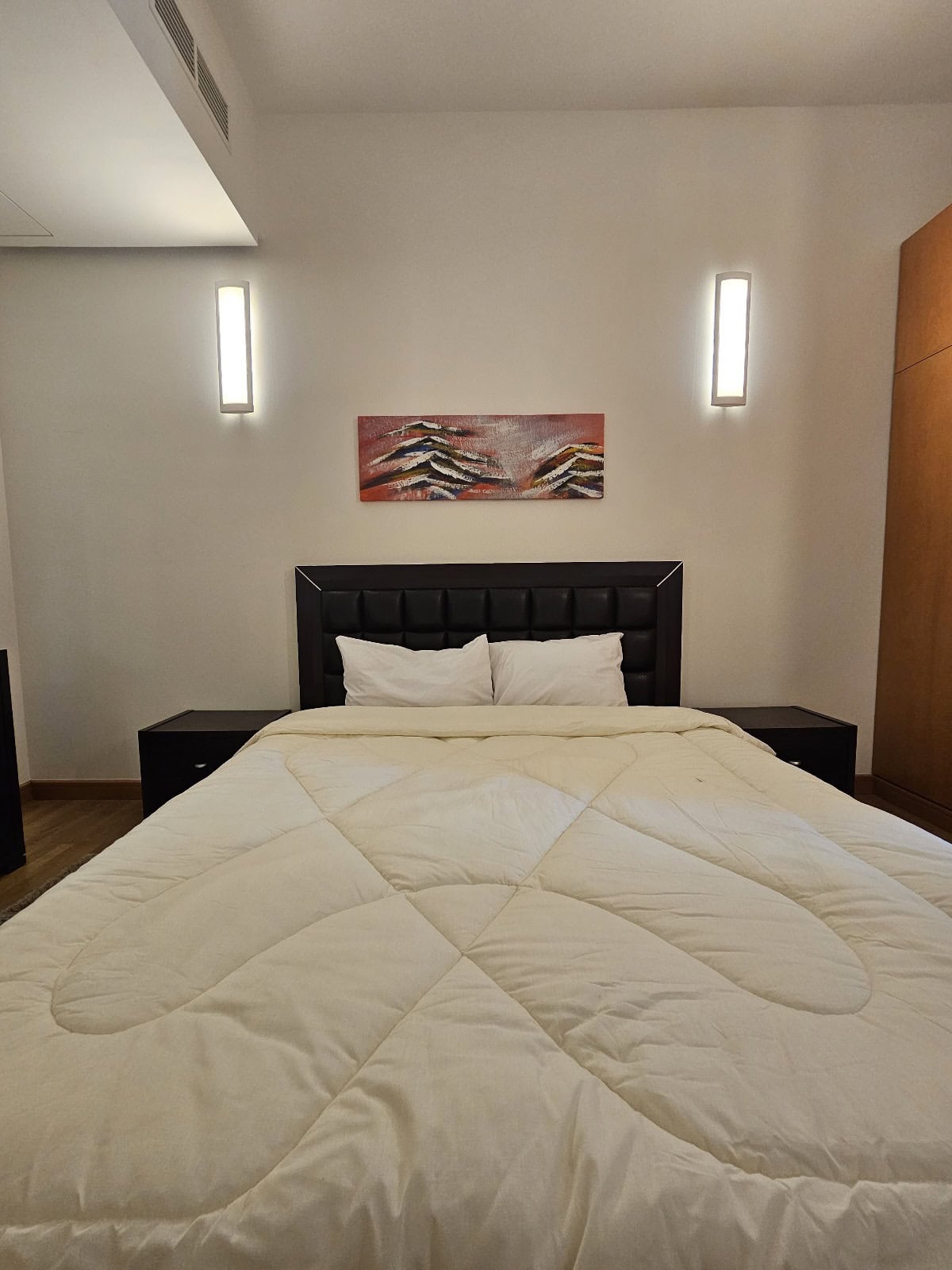 A neatly made bed with a beige quilt, black headboard, two pillows, and two side tables with wall-mounted lights above graces this flat for rent on Reef Island. A colorful abstract painting hangs on the wall, adding a touch of vibrancy to the serene bedroom.