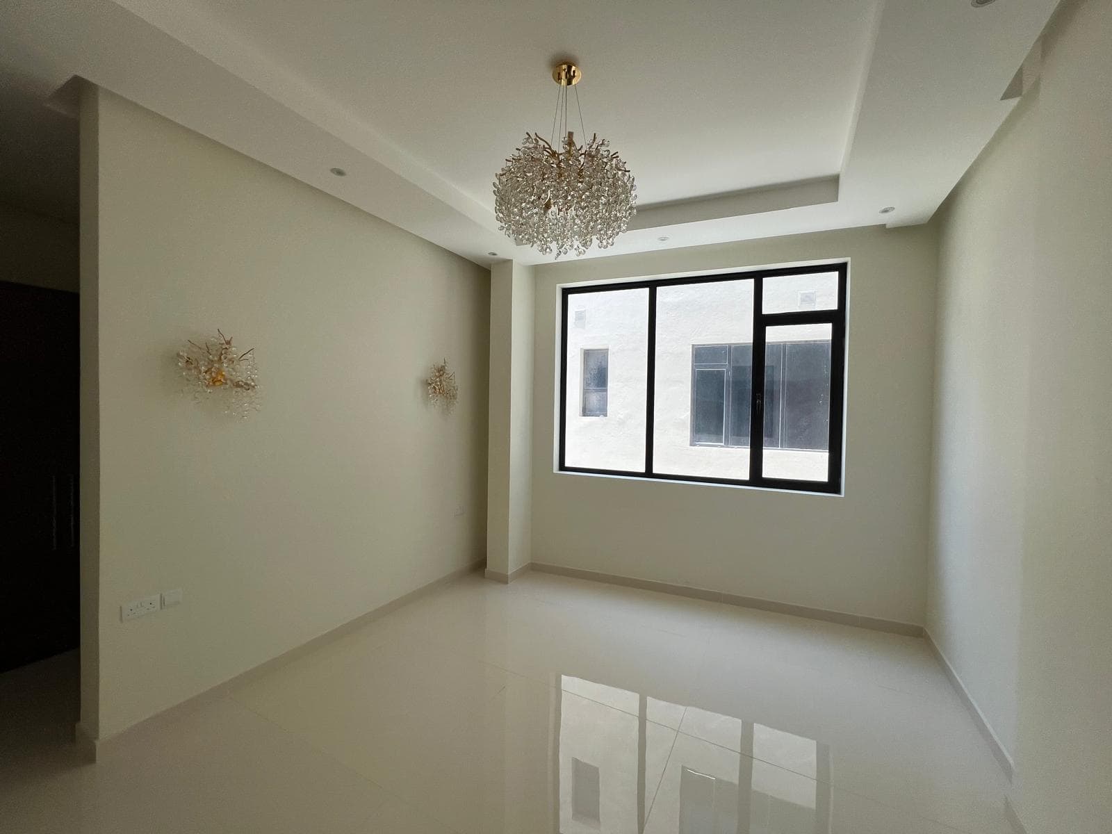 A modern, empty room with beige walls, a tiled floor, a large rectangular window, a chandelier, and two wall-mounted light fixtures.