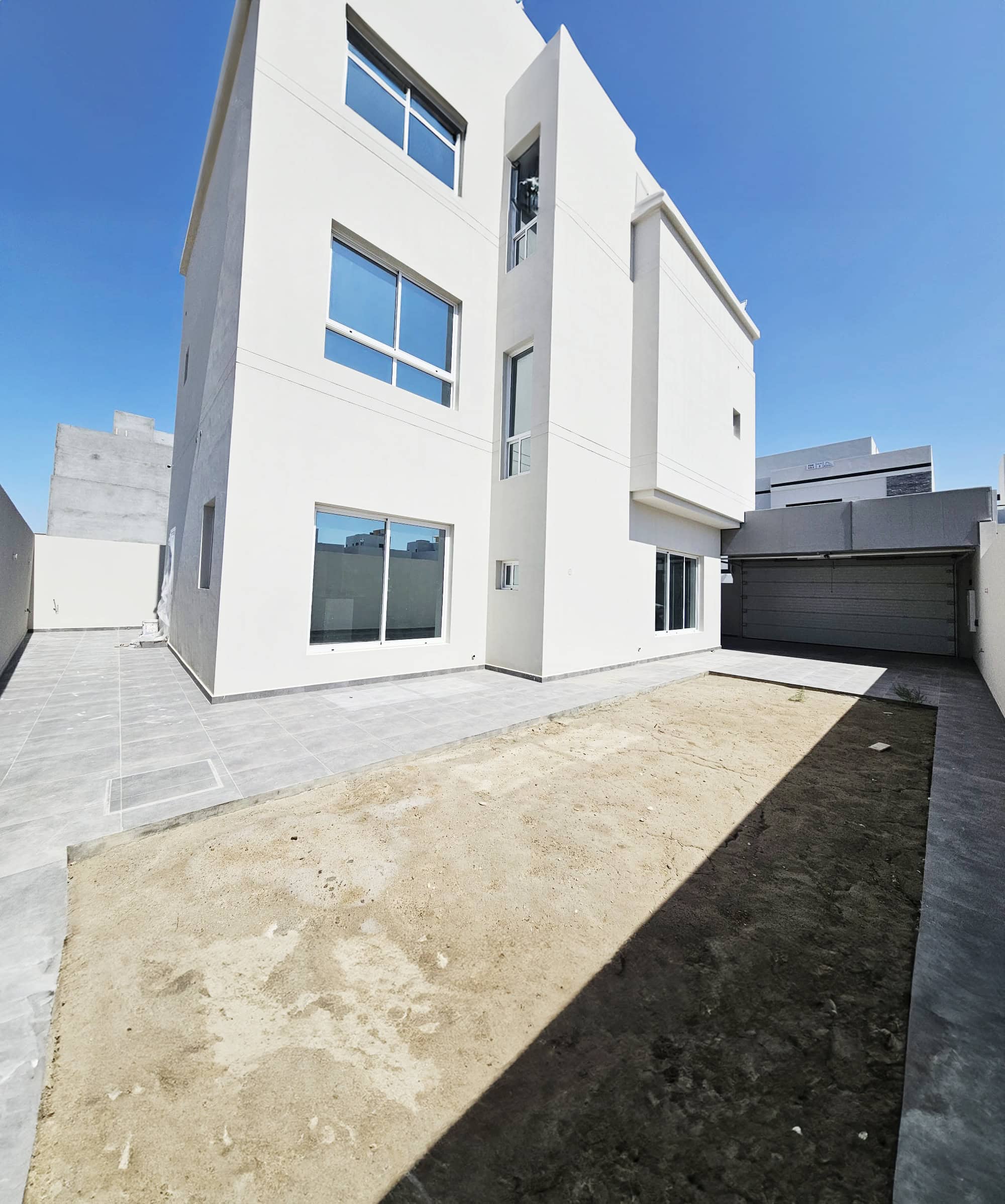 A modern, three-story white building with large windows stands proudly under a clear blue sky. The empty sandy yard and adjacent tiled pathways create a serene environment, showcasing the impeccable design that's akin to an auto draft of architectural brilliance.