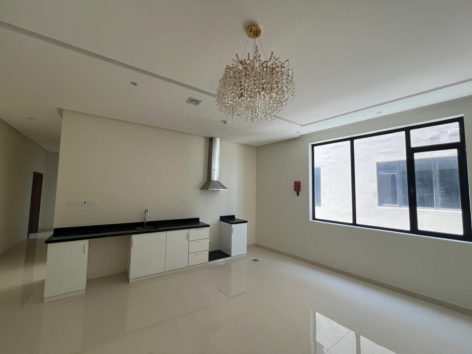 A modern, unfurnished kitchen and living area with white walls, black countertops, a stainless steel range hood, large window, and a chandelier. The floor is tiled in white.