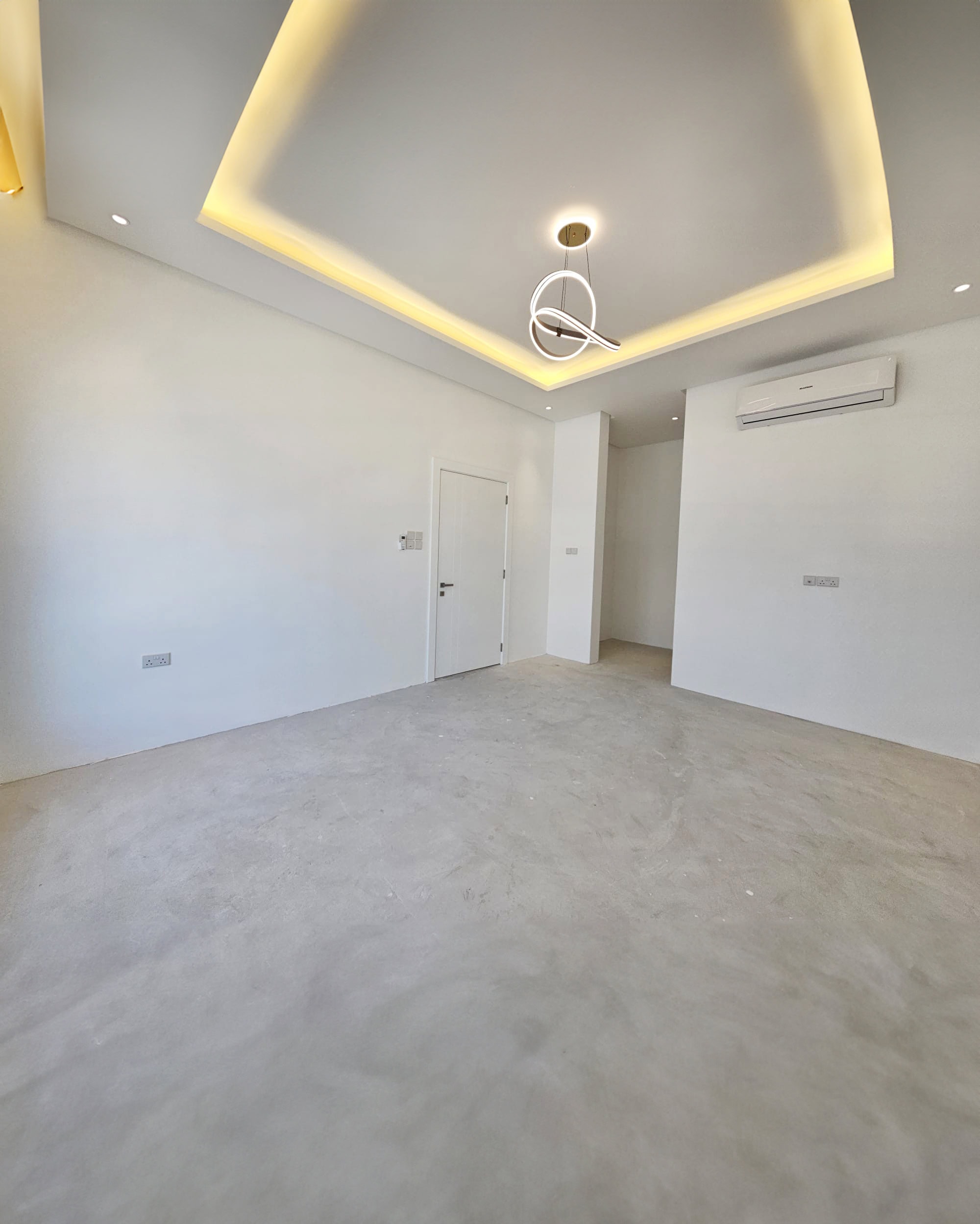 A vacant room with white walls, an unfinished concrete floor, a modern ceiling light fixture, and an air conditioning unit mounted on the wall. There are two doorways and recessed lighting on the ceiling—an inviting space in the luxurious villa at Saraya 1, now available for sale.