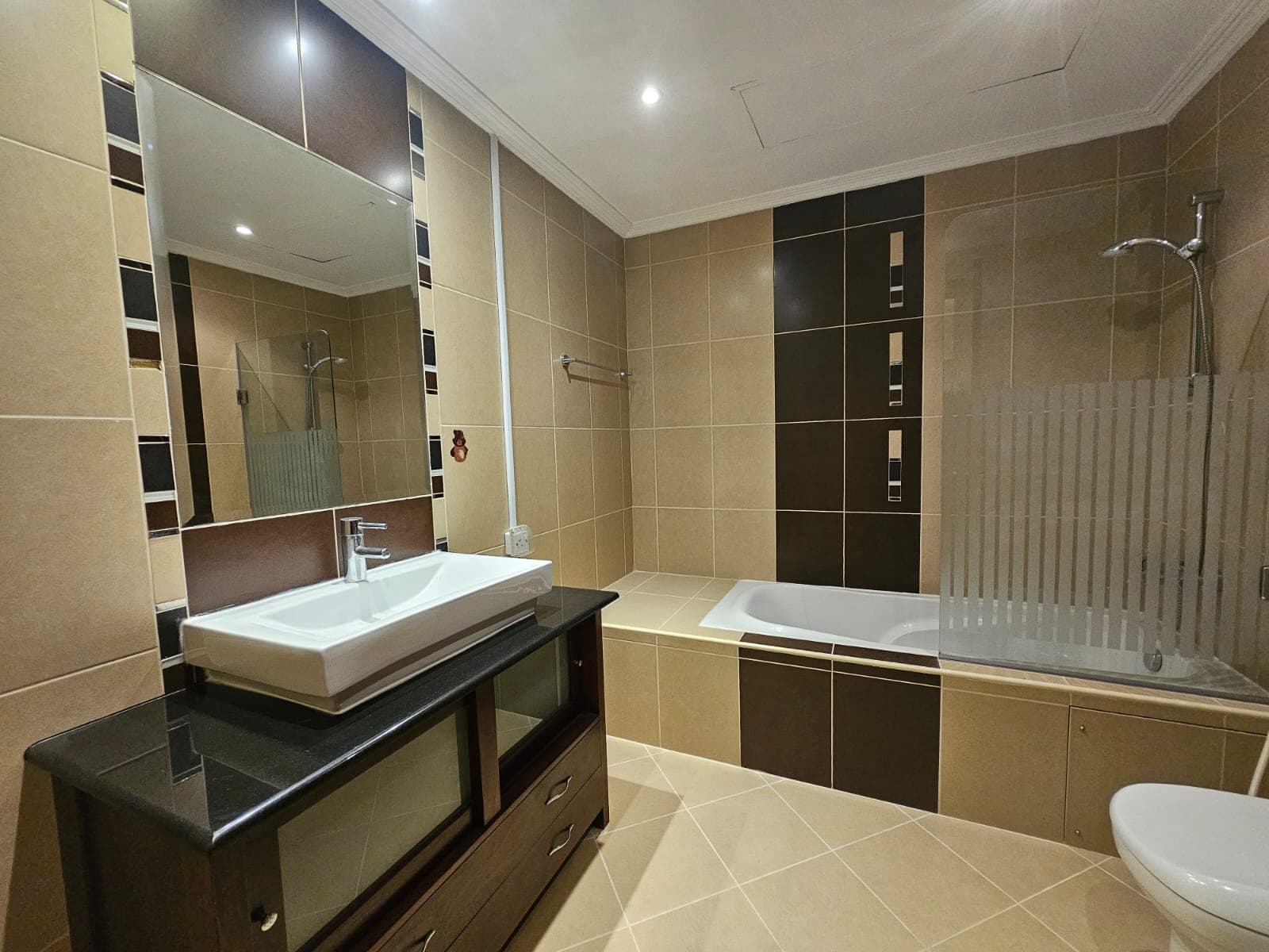 This flat for rent in Juffair features a modern bathroom with beige and dark brown tiles, a bathtub with a glass partition, a white sink on a black countertop, a wall mirror, and a toilet.