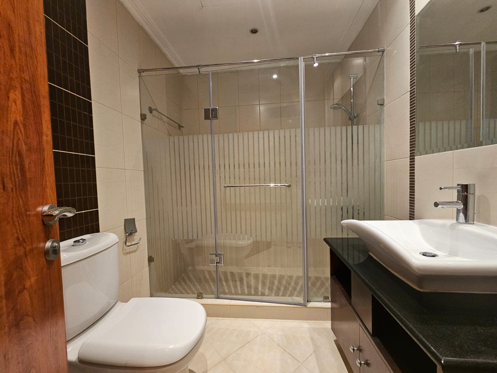 A modern bathroom in a Juffair flat for rent, featuring a glass-enclosed shower, a white toilet, a stylish sink on a black countertop, and beige tiles on the floor and walls.