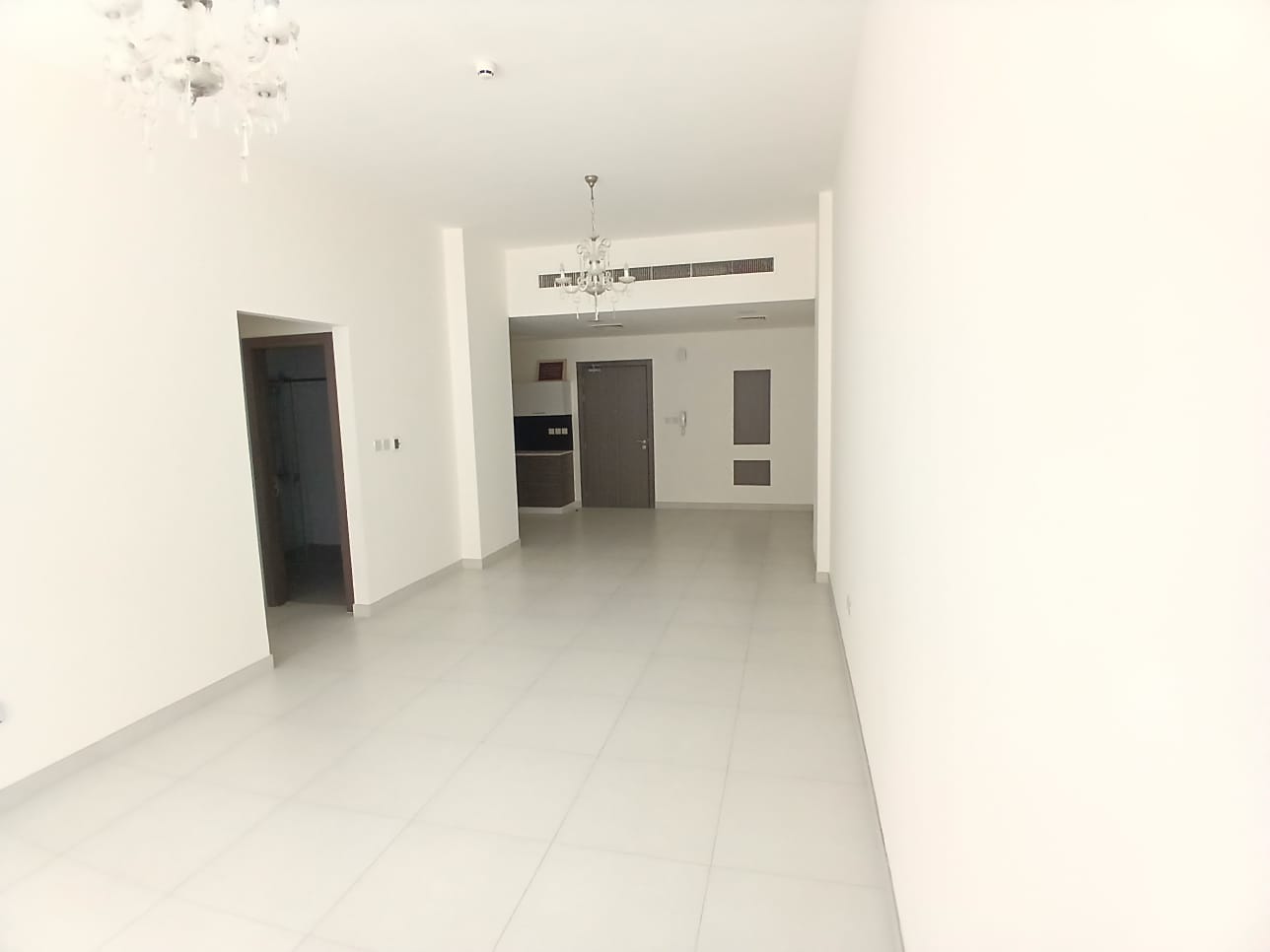 A spacious, empty room with white walls and tiled floor. The room features a chandelier, a door, and hallway entrances on the back wall.