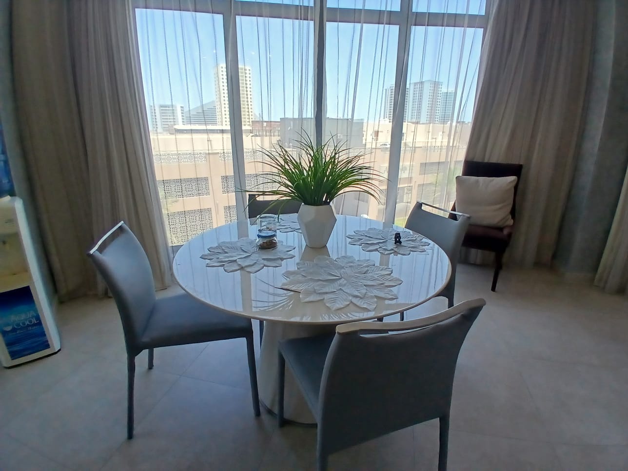 A round dining table with a white centerpiece and four chairs is set next to tall windows with sheer curtains, overlooking a stunning Amwaj cityscape. This chic setup is perfect for anyone seeking a flat for rent with a view of the skyline.