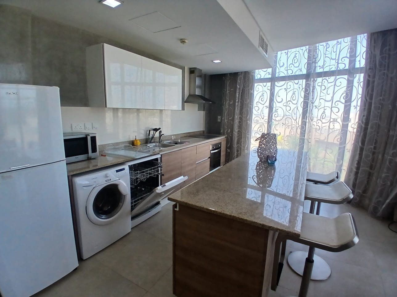 Modern kitchen with white cabinets, a refrigerator, a washer, an oven, a stovetop, and a dishwasher. A granite island with two stools is in the center, next to a large window with sheer curtains. This Amwaj flat for rent offers elegance and functionality in every detail.