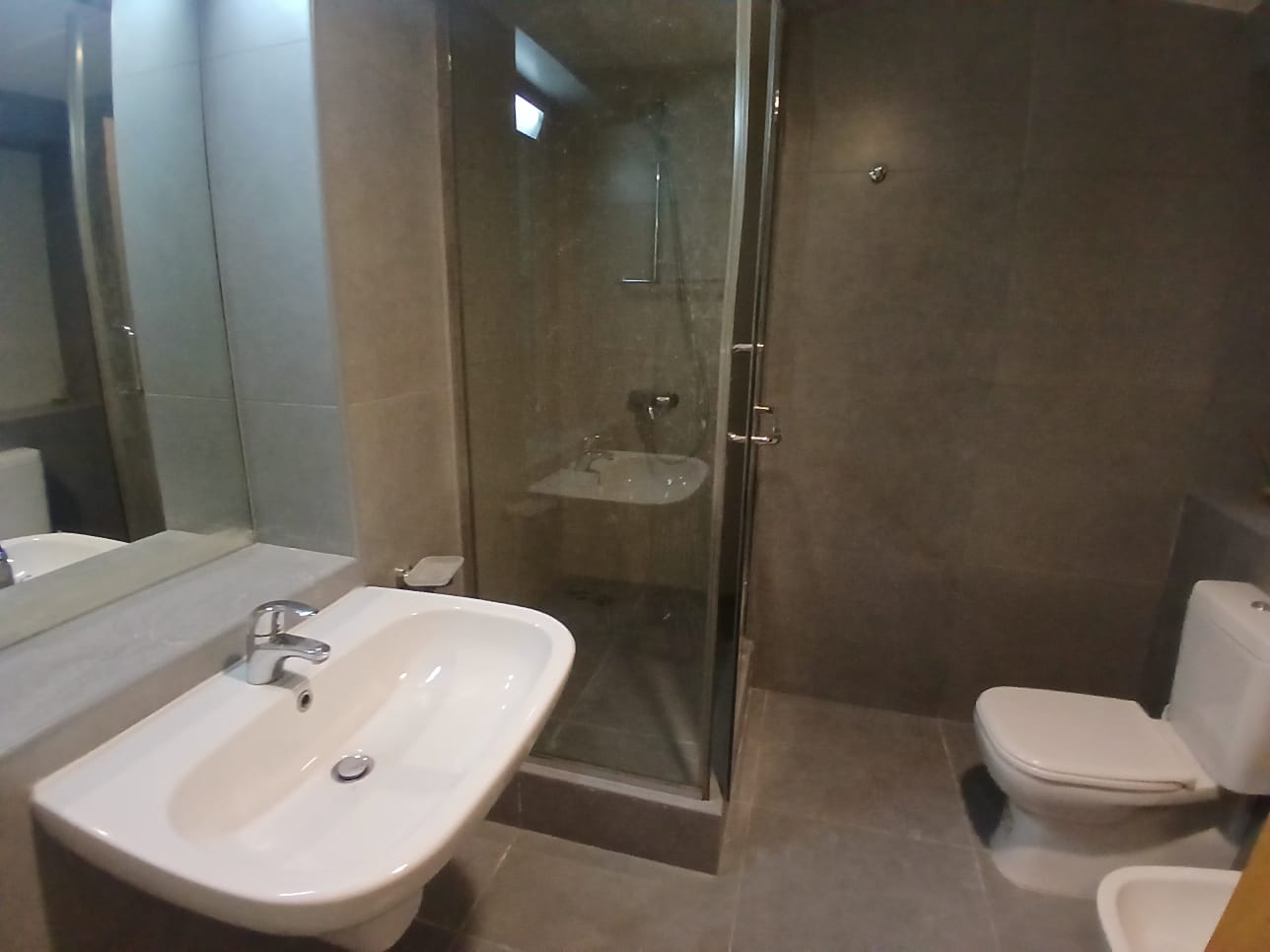 A flat for rent in Amwaj features a modern bathroom with a sink, a mirrored wall, a glass-enclosed shower, and a toilet. The fixtures and tiles are all in neutral tones.