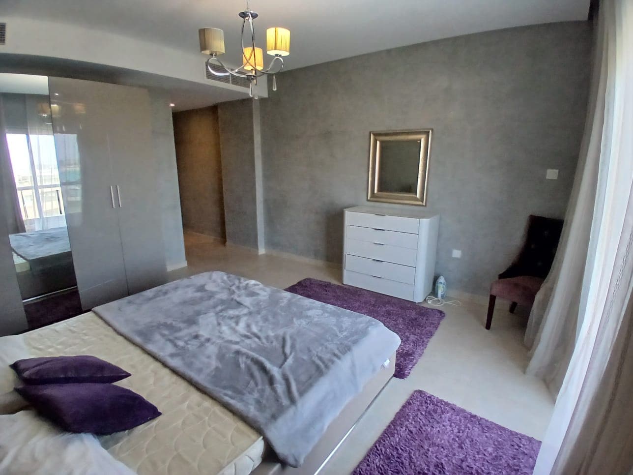 A modern bedroom in an Amwaj flat for rent featuring a bed with gray and purple pillows, a matching throw, a chest of drawers with a mirror above it, a chair, and a closet with mirrored doors. Soft lighting filters through curtains.