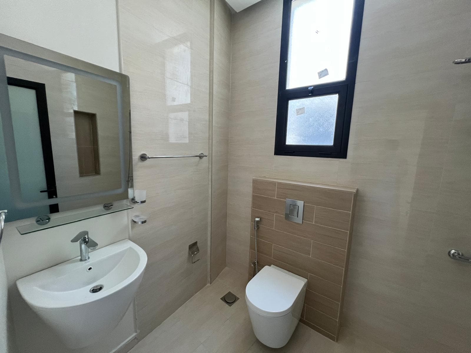 A small, modern bathroom with a white sink, a wall-mounted toilet, a frosted window, and beige tiled walls and floors. A mirror hangs above the sink.