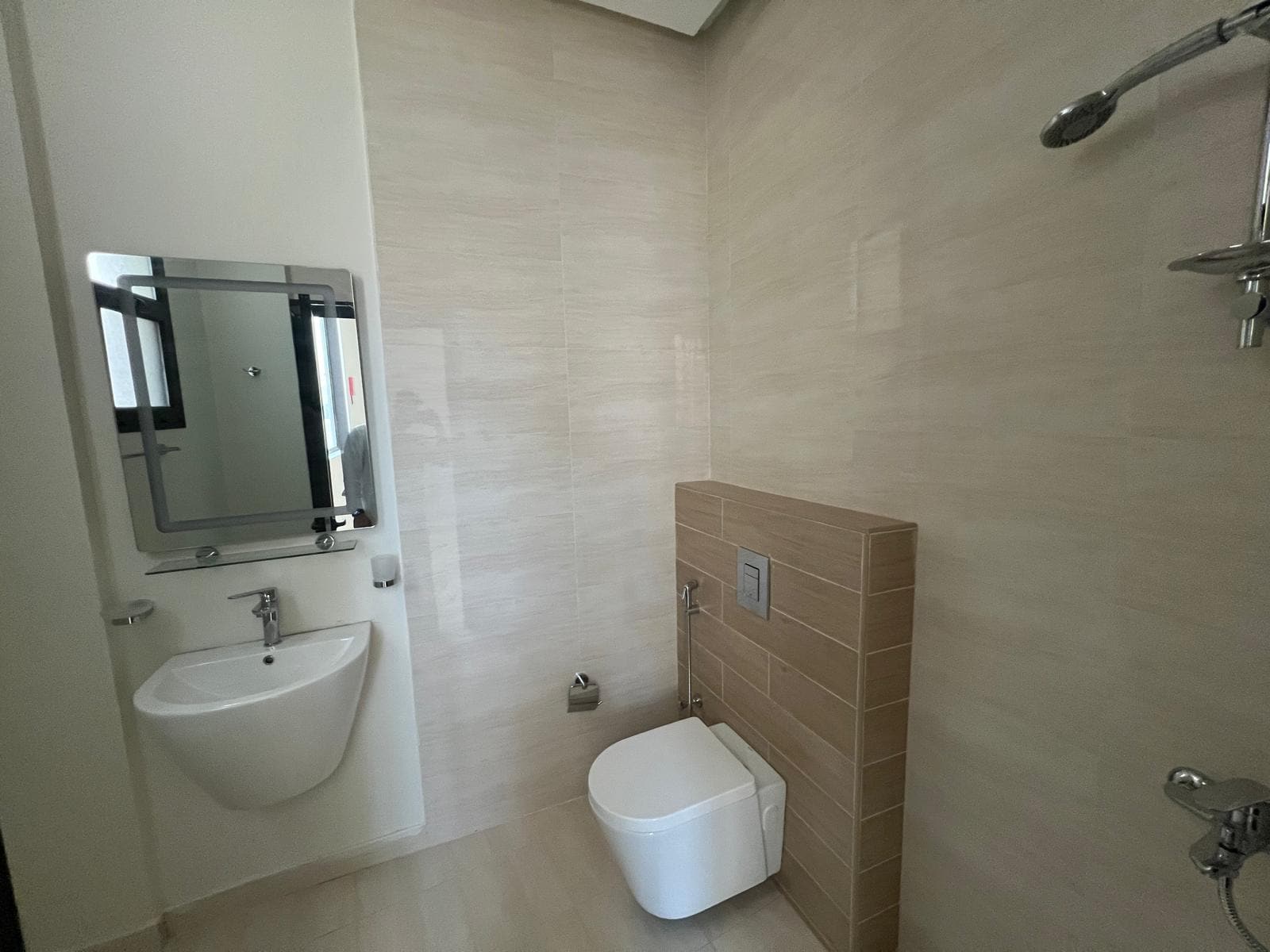 A modern bathroom with beige tiles, a wall-mounted toilet, a sink with a mirror above it, and a shower on the right side.