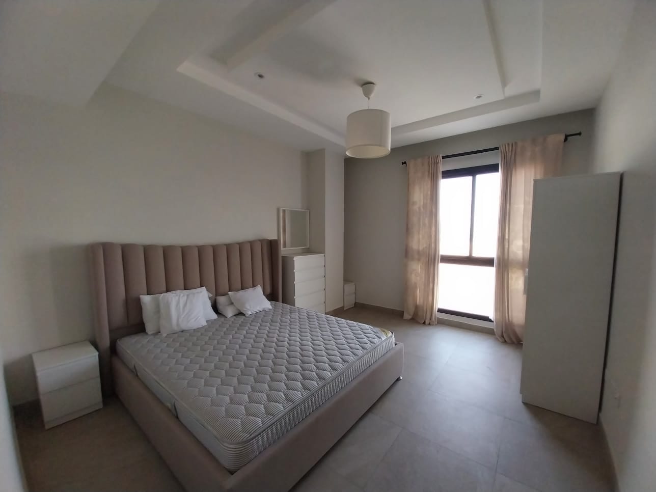 A minimalist bedroom in a flat for rent featuring a large bed with a padded headboard, two side tables, a chest of drawers with a mirror, and a window with curtains allowing natural light to enter the room.