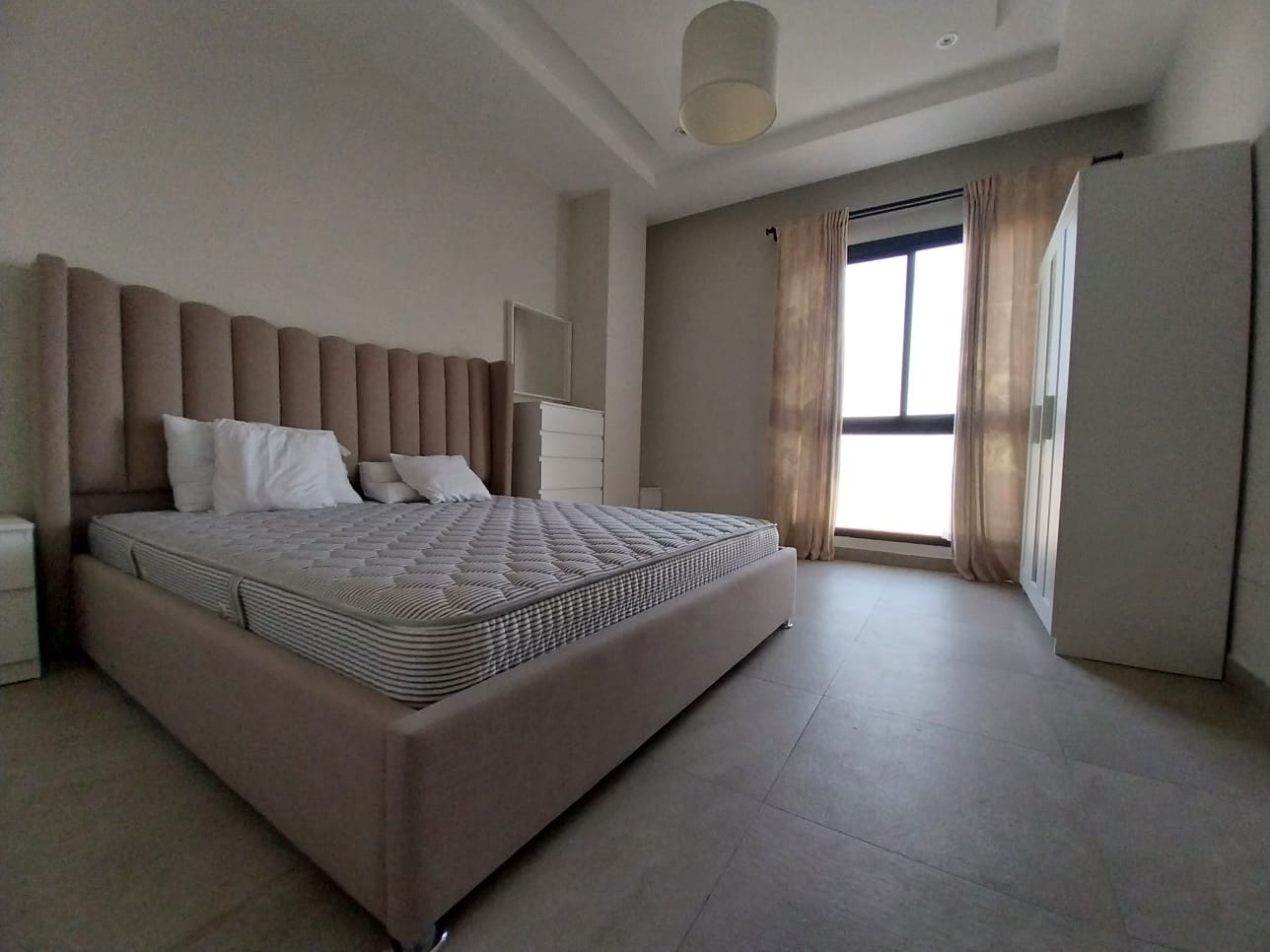 A minimalist bedroom in a flat for rent in Hidd features a large bed with two pillows, a soft pink padded headboard, and a wardrobe near a window with sheer curtains. The walls are plain and the room is well-lit, creating an inviting space for relaxation.