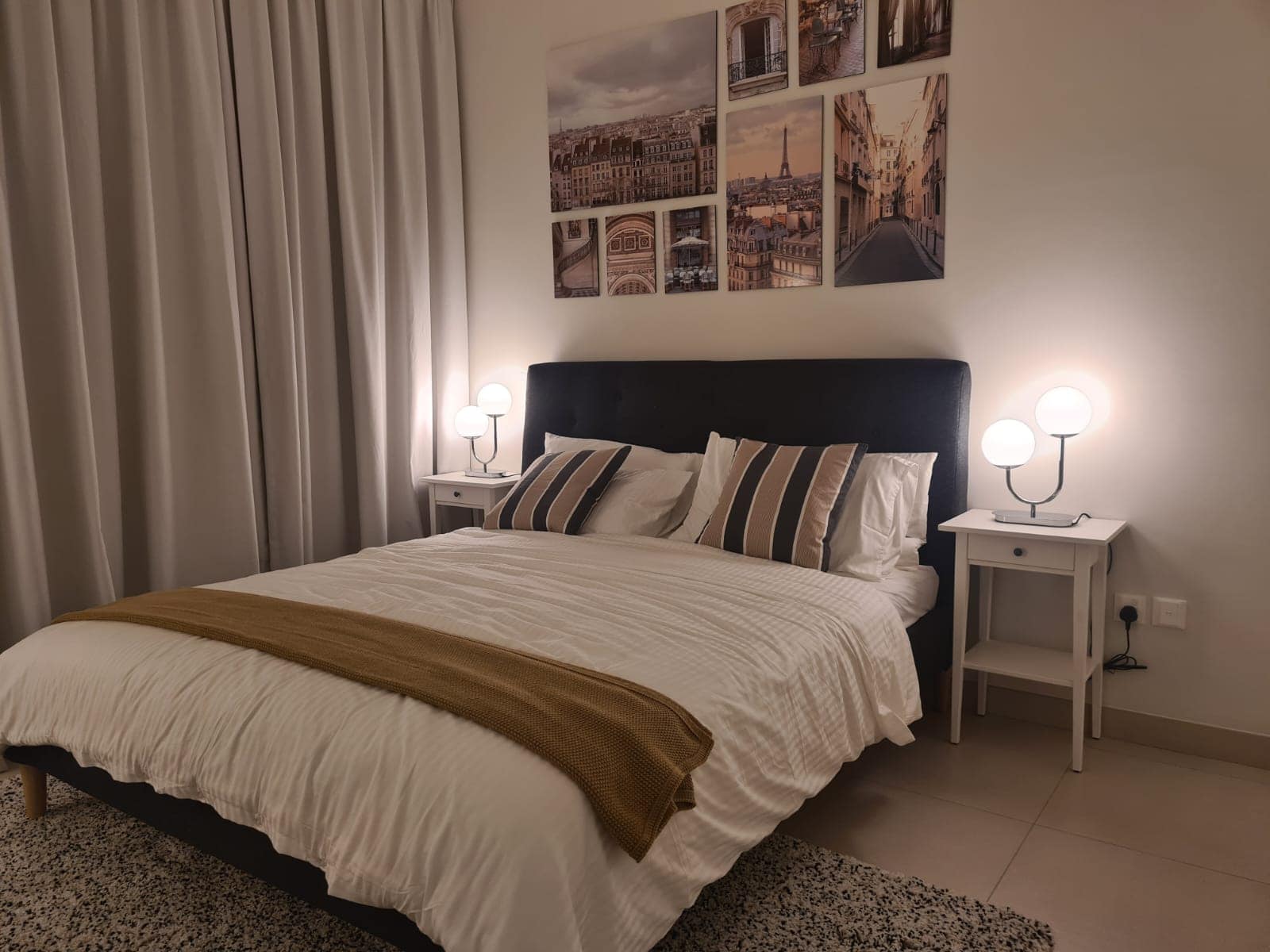 A neatly made bed with white and gray bedding, accented by striped pillows and a brown throw blanket, is showcased in this flat for sale in Diyar Al Muharraq. Two bedside tables with modern lamps are positioned on either side, and framed photos adorn the wall above.