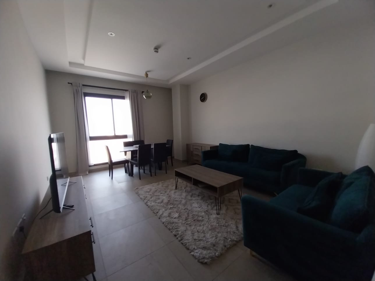 A flat for rent in Hidd features a living room with a dark teal sofa set, a wooden coffee table, a TV on a stand, and a dining table with chairs. Light curtains dress the window, while tiled floors and a cozy rug complete the setup.