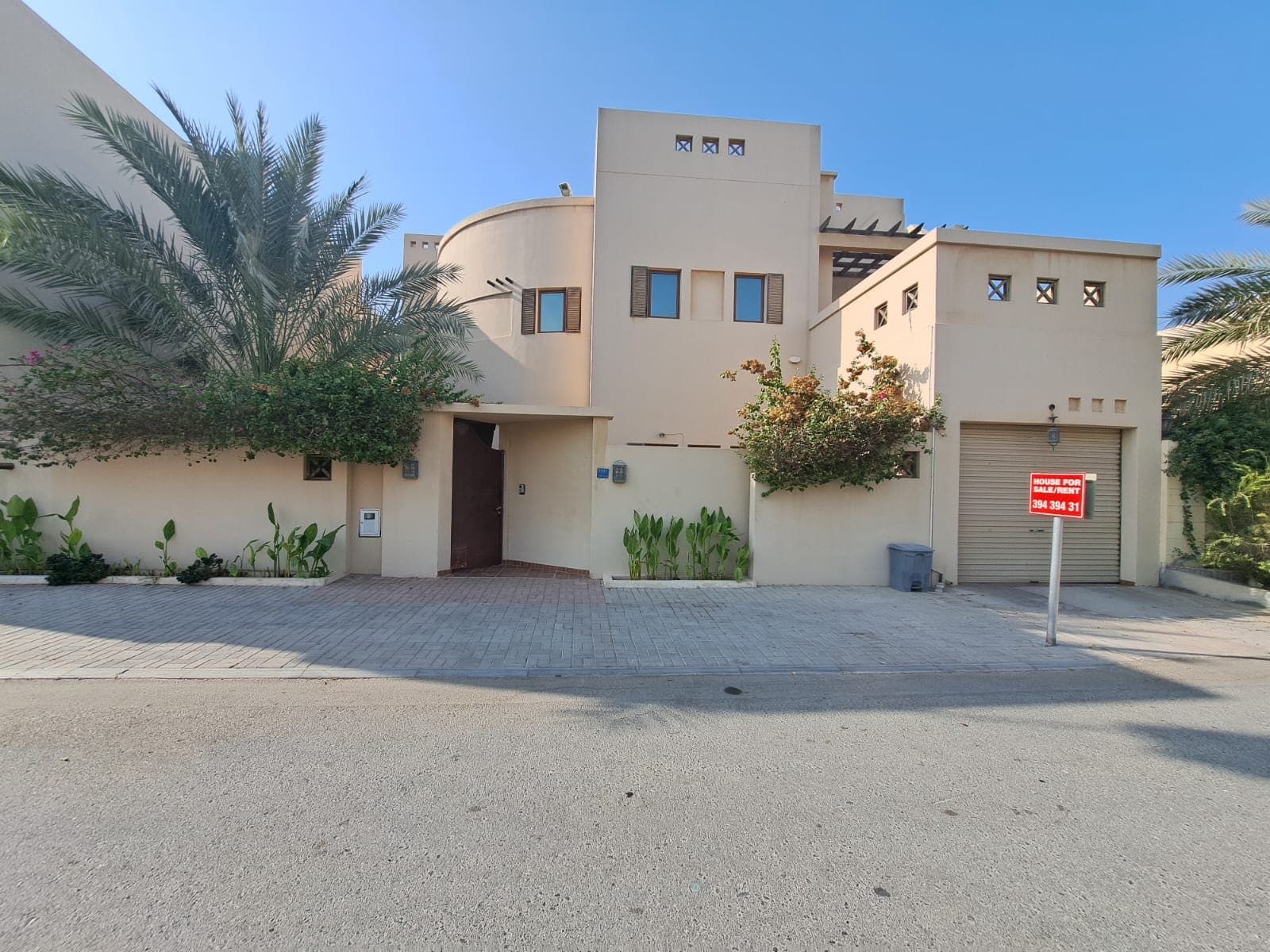 A luxurious villa in Saraya 1, this modern tan two-story house features small windows, a gray garage door, and is surrounded by various plants. With a "For Sale" sign prominently displayed, the driveway and street are also visible.