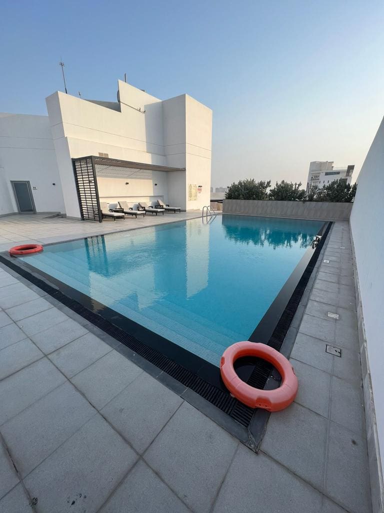 A rooftop swimming pool with clear water and several lounge chairs in the background, available for rent in Juffair. Two orange life rings are placed on the pool deck. Trees and buildings are visible in the distance, adding to the serene ambiance of this luxurious apartment amenity.